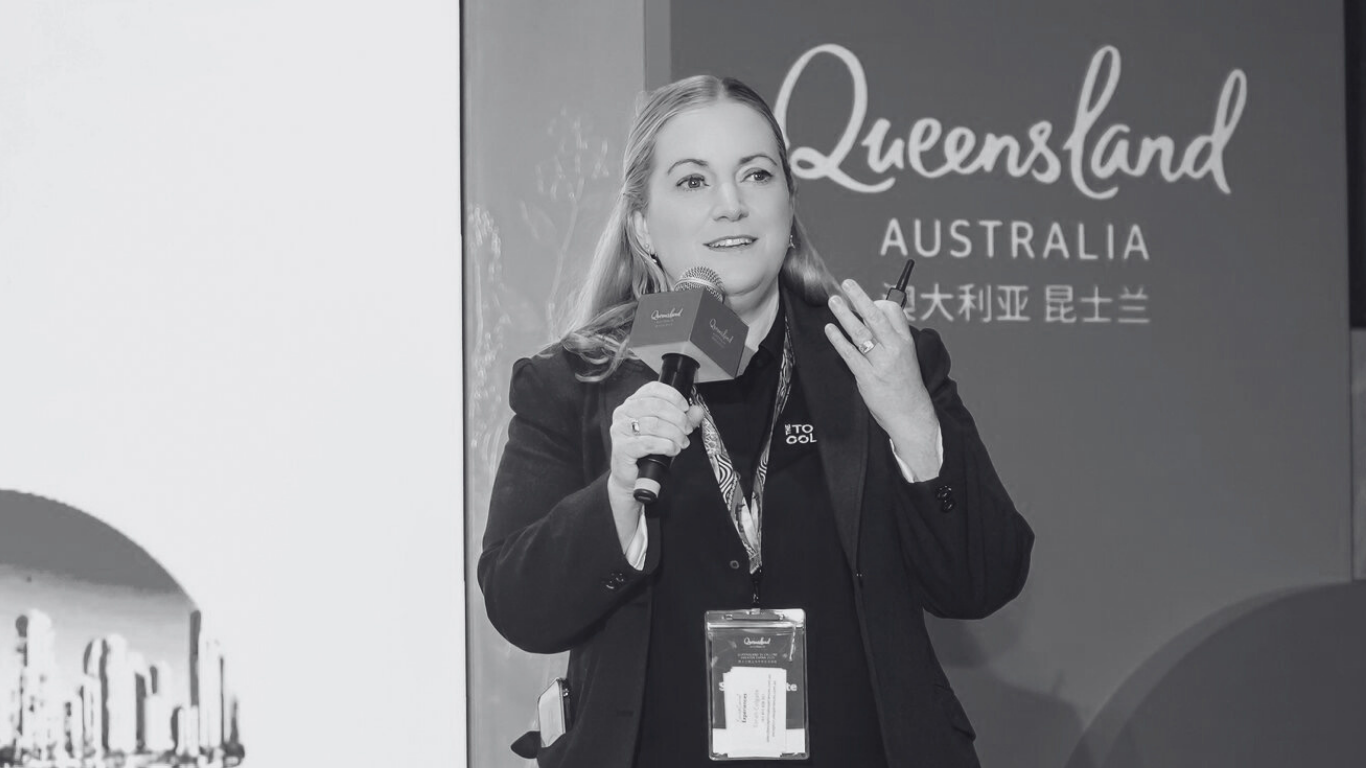 A woman speaking into a microphone at an event with a 'Queensland Australia' banner in the background.