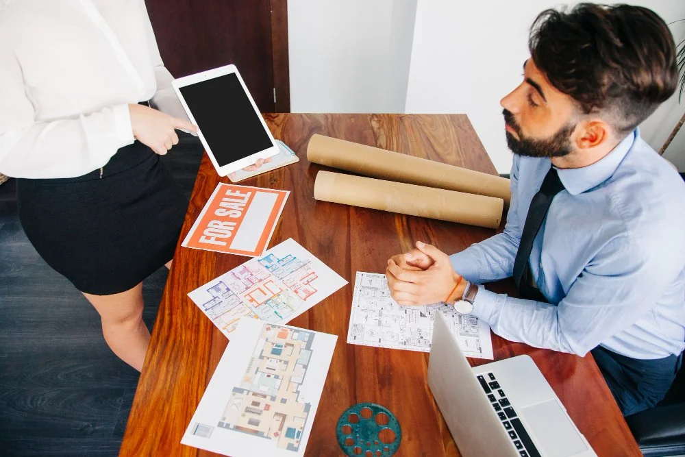 Real estate agent showing a tablet to a couple sitting at a wooden table with house plans, a 'For Sale' sign, and a laptop.