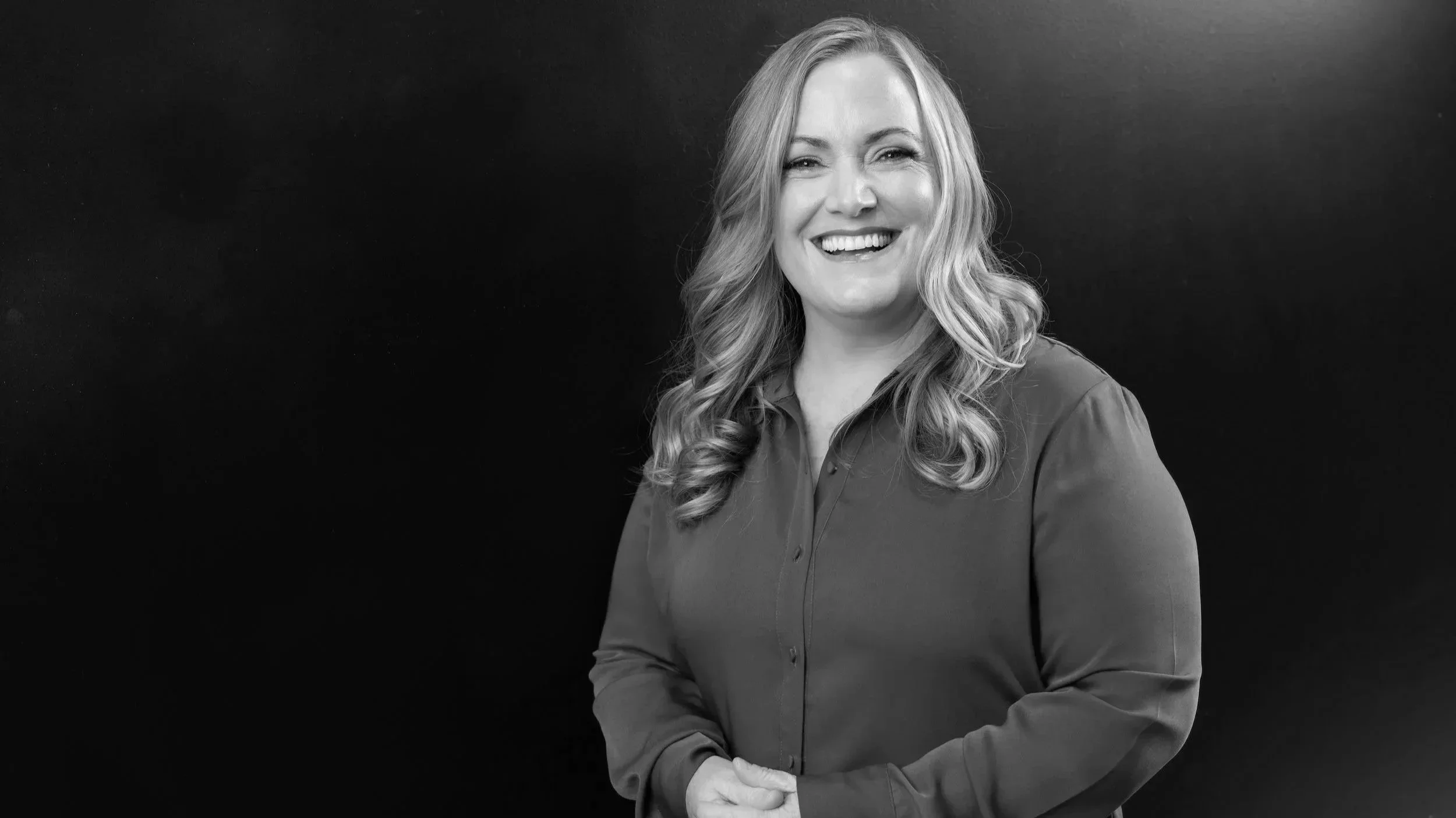A Black and white portrait of a woman with long wavy hair, smiling and looking at the camera, wearing a button-up shirt against a dark background.