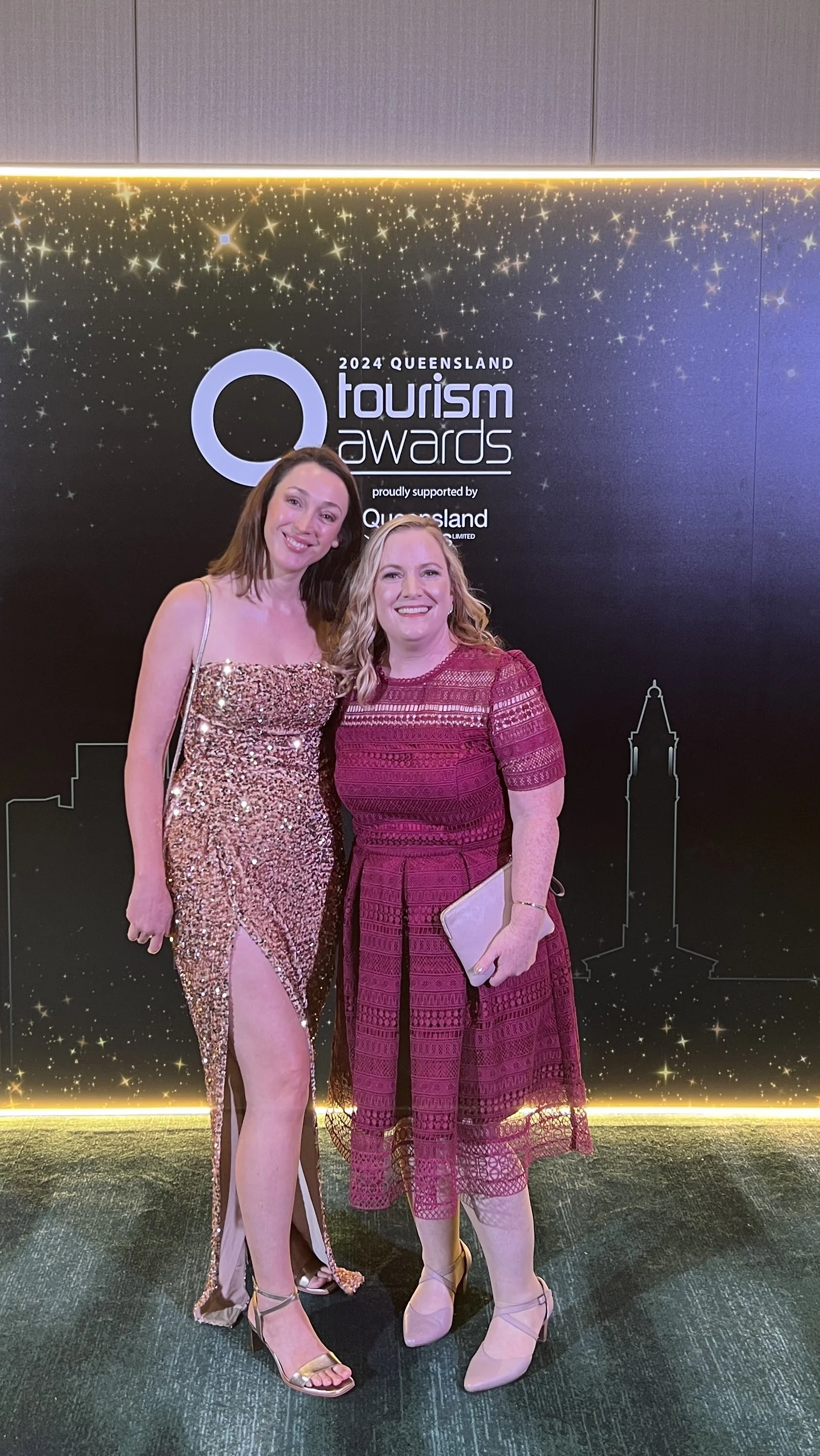 Two women in elegant dresses standing in front of a backdrop for the 2024 Queensland Tourism Awards, smiling at the camera.