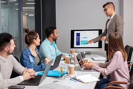 Business meeting in a conference room with five people, one standing and presenting financial charts on a screen, others seated with laptops and notebooks.