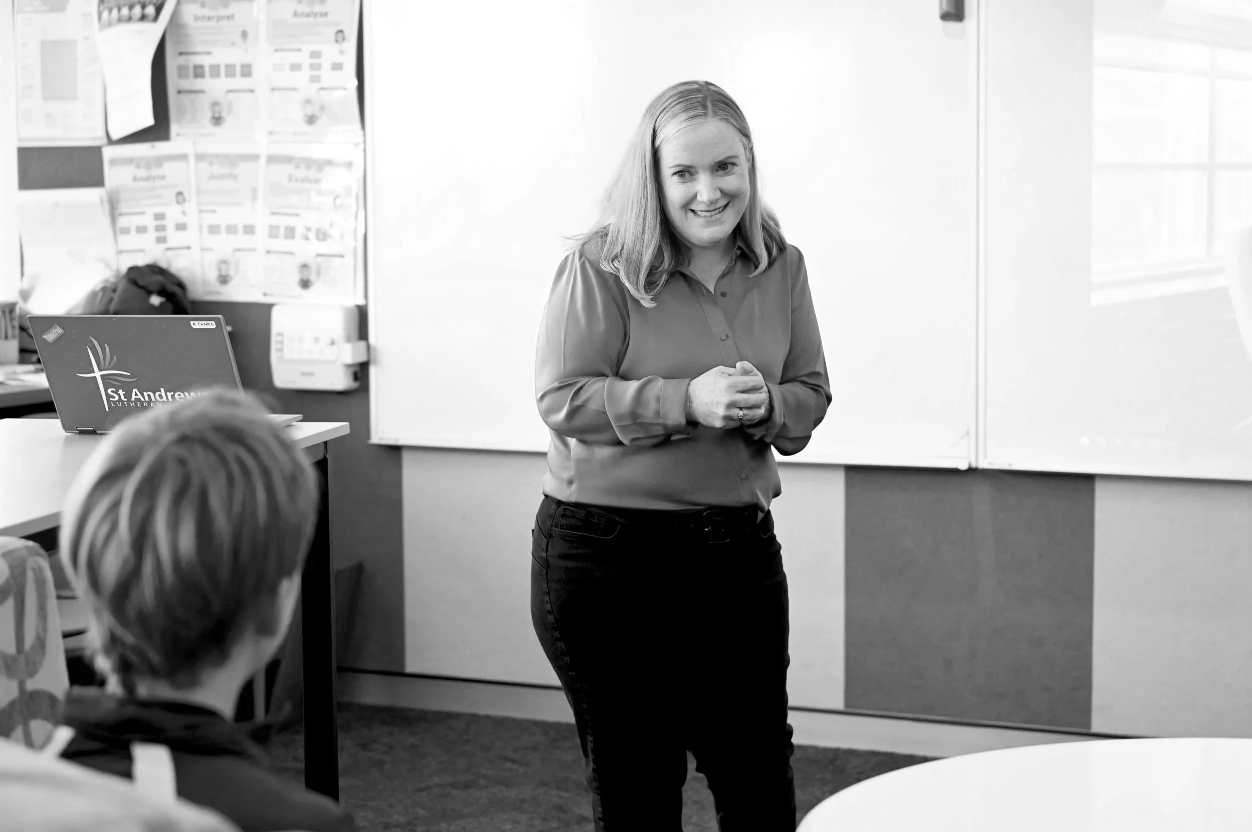 Sarah Colgate smiling and standing in front of a classroom whiteboard, speaking to a seated person with short hair.
