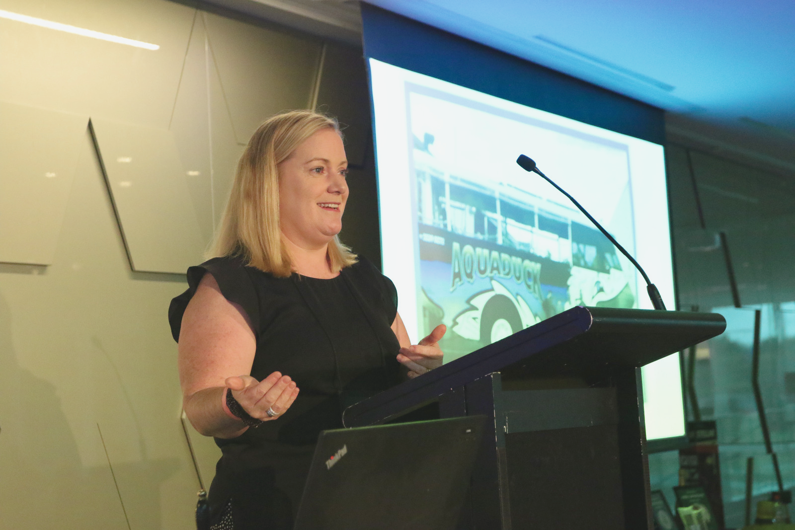 A woman giving a presentation at a podium with a screen displaying a boat with the word 'Aquaduck' in the background.