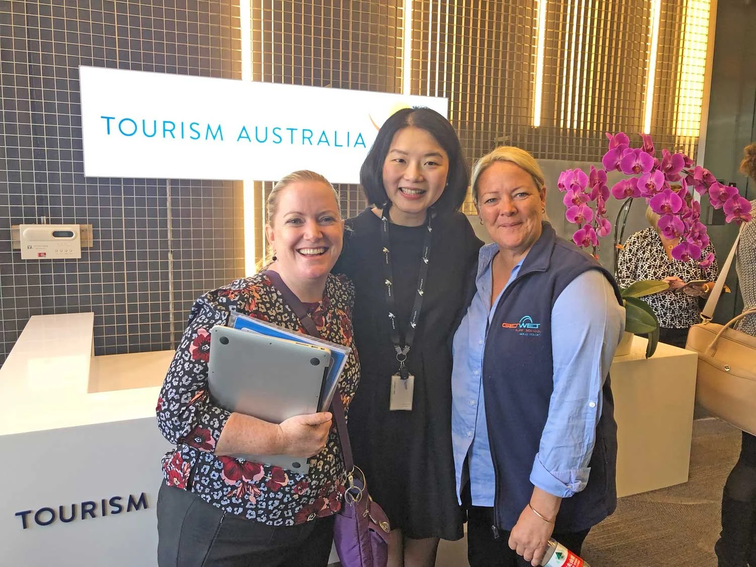 Three women smiling posing together at a tourism Australia event with a sign and purple orchids in the background.