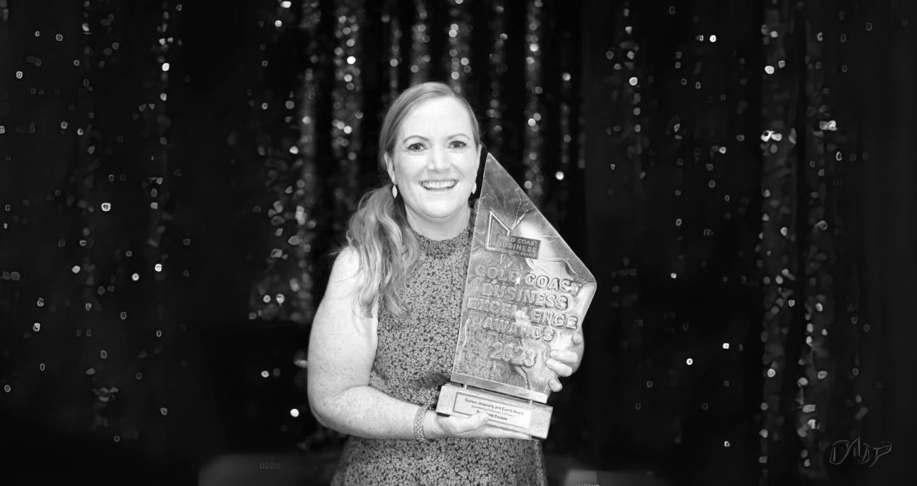 Sarah Colgate with long wavy hair wearing a floral dress, holding an award trophy at a celebration event with a sparkly backdrop, smiling at the camera.