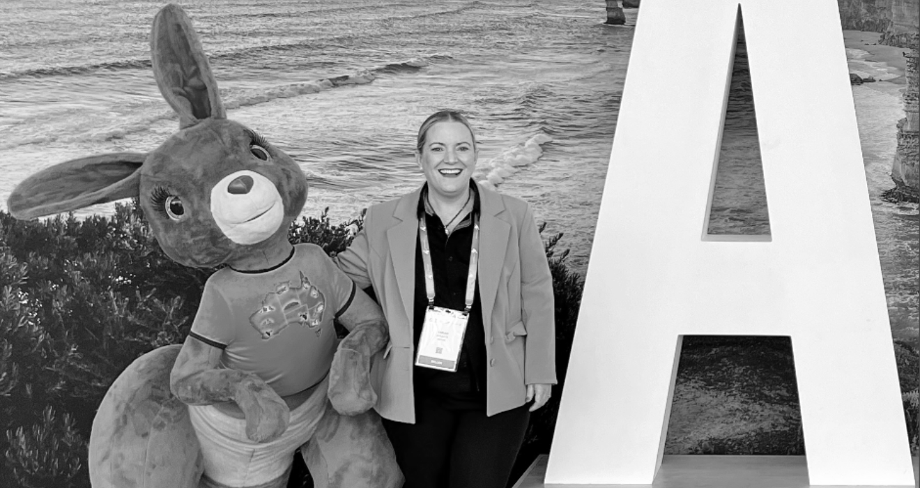 A woman smiling while posing with a person in a large Easter bunny costume. The background features a large letter 'A' and a coastal scene with rocks and water.