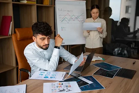 Man working on laptop with documents, woman presenting charts on a flip chart in a modern office conference room.