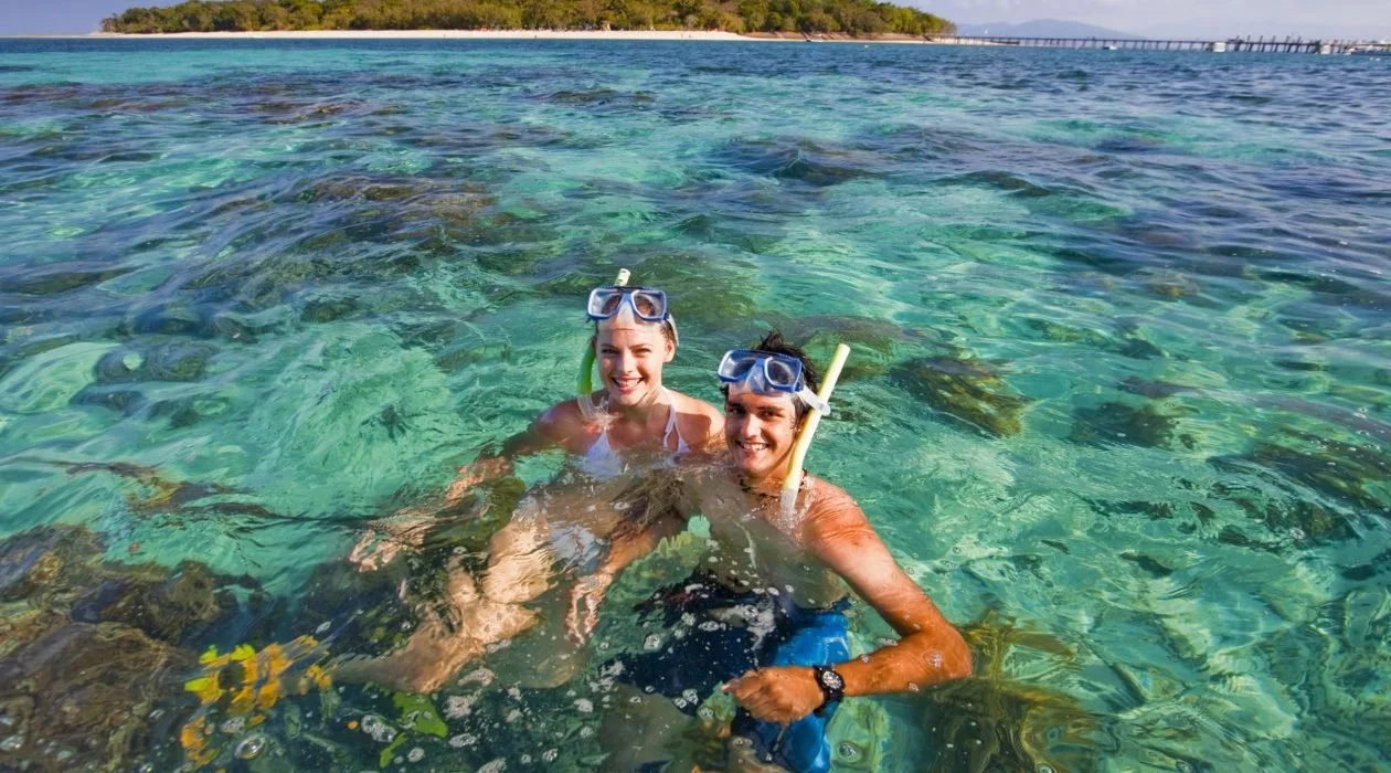 Two women in snorkeling gear smiling in clear turquoise ocean water near a rocky shore with a distant tree-lined island and bridge in the background.