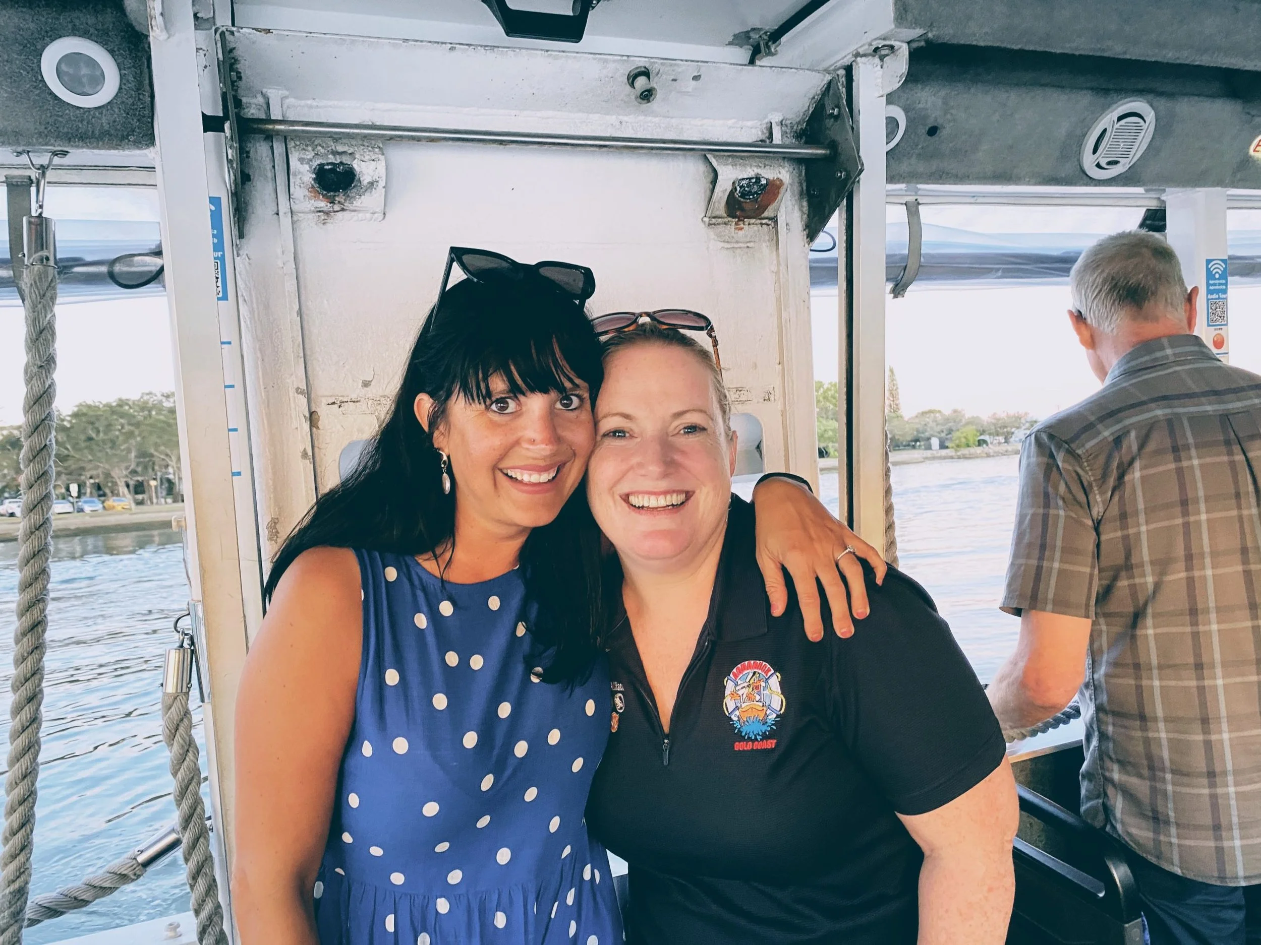 Two women smiling and hugging on a boat with water and trees in the background, one wearing a blue polka dot dress and the other a black polo shirt.