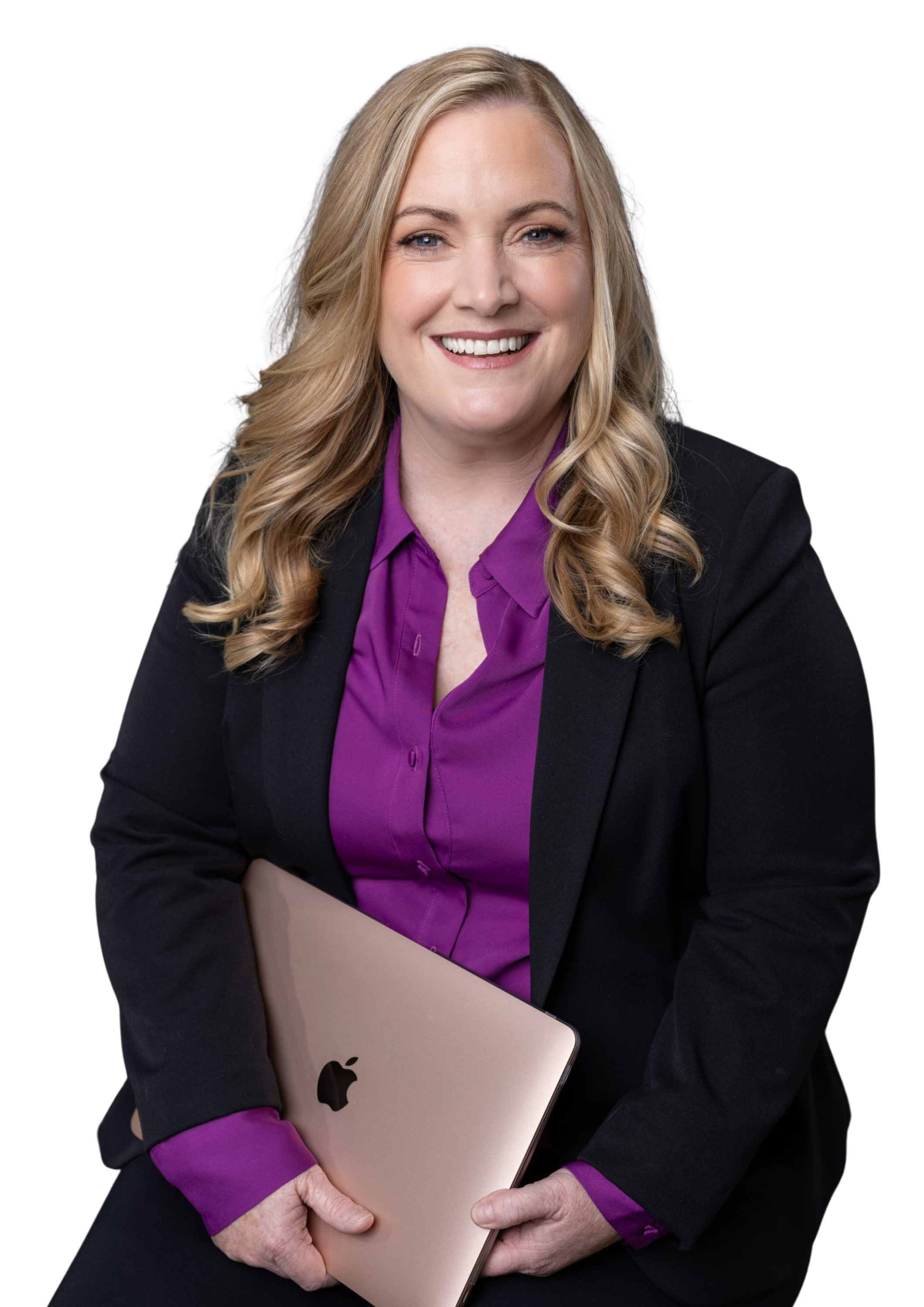 A smiling woman with blonde wavy hair, wearing a black blazer and purple blouse, holding a closed rose gold MacBook, against a white background.