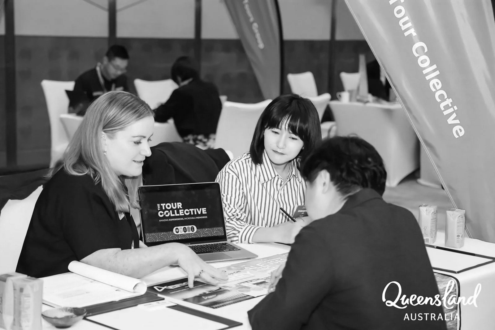 Three people sitting at a table engaging in a discussion or interview, with promotional materials and laptops on the table, at an event in Queensland, Australia.