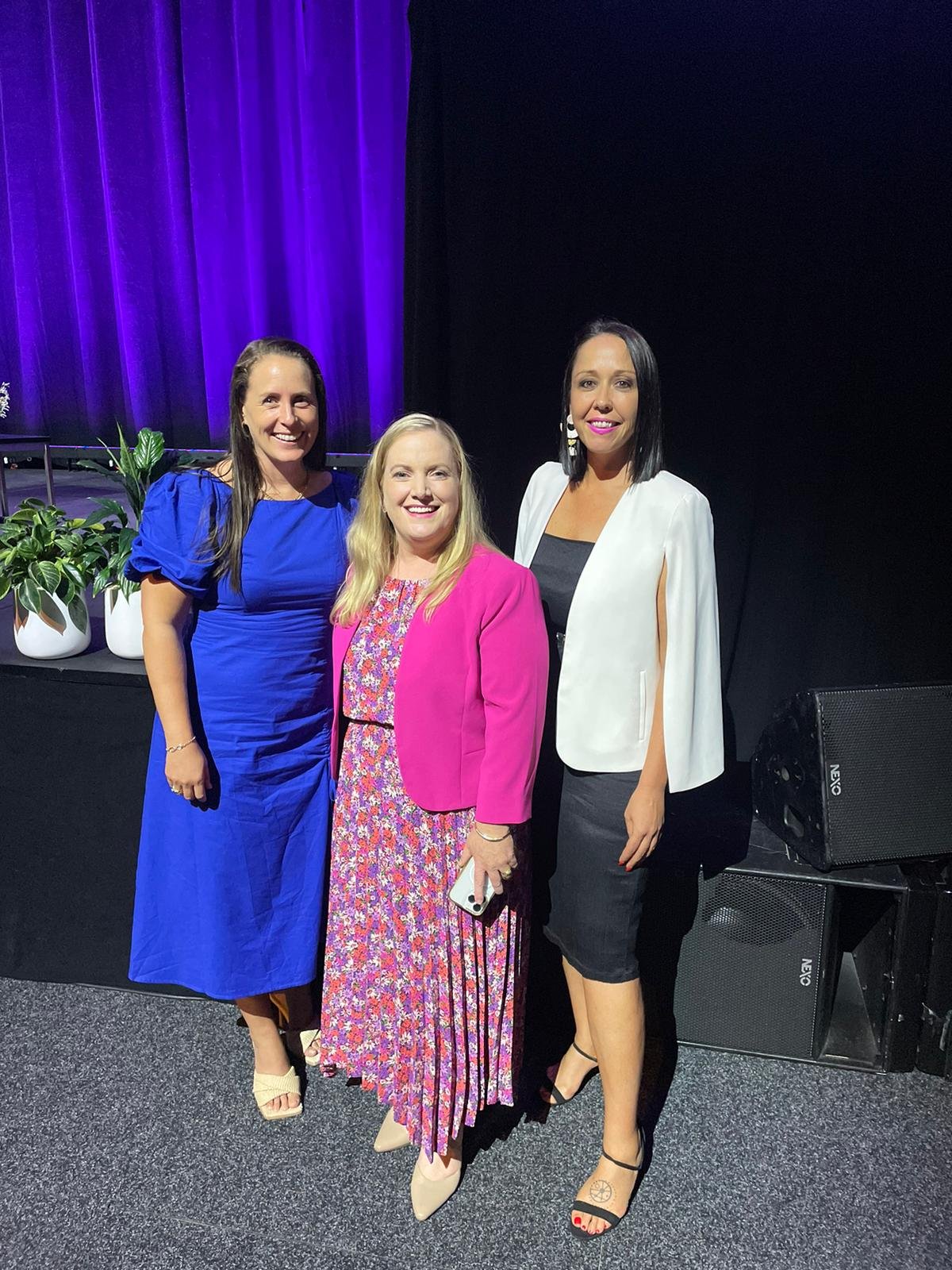 Three women standing together on a stage with purple curtains, posing for a photo, with potted plants in the background.