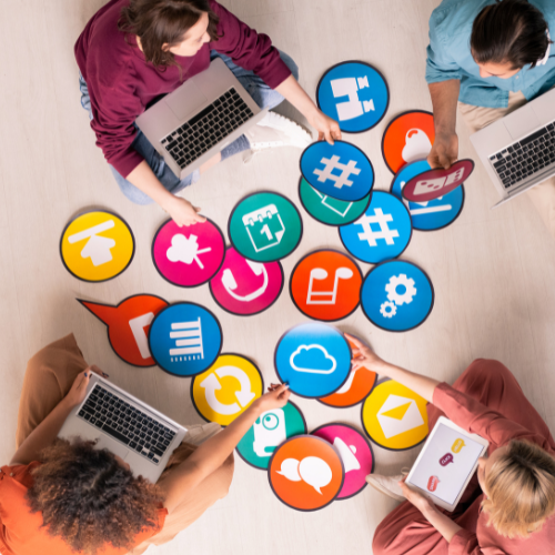 Four people sitting around a table with laptops and colorful icon cutouts representing social media and communication symbols.