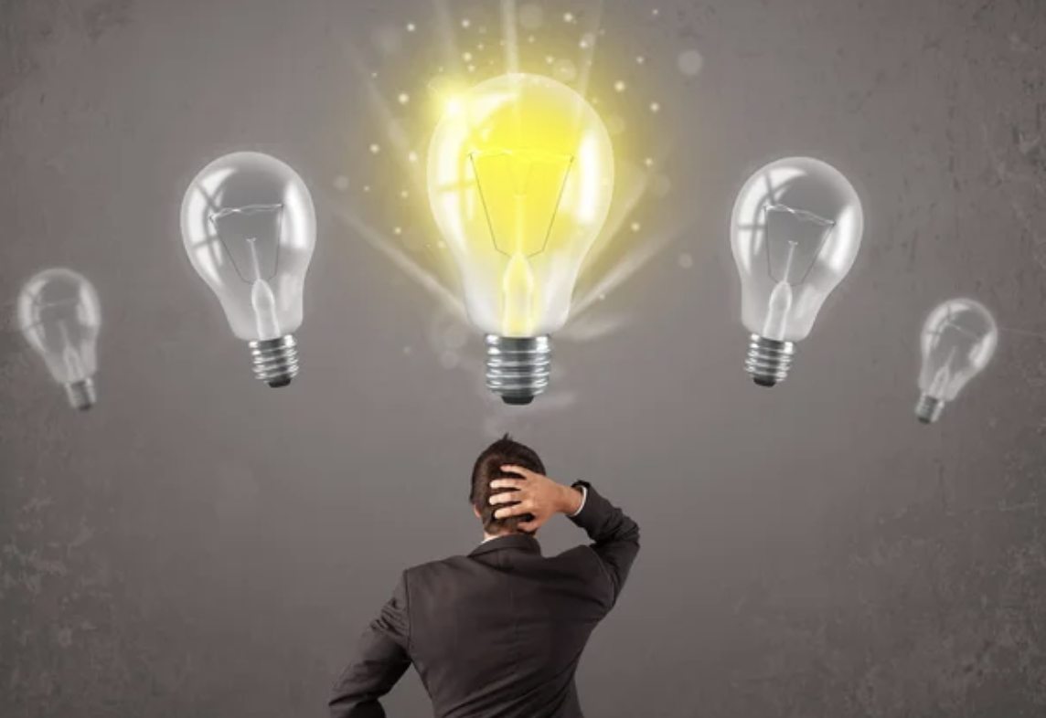 A man in a suit standing with his back to the camera, holding his head, surrounded by floating light bulbs, one of which is glowing yellow.