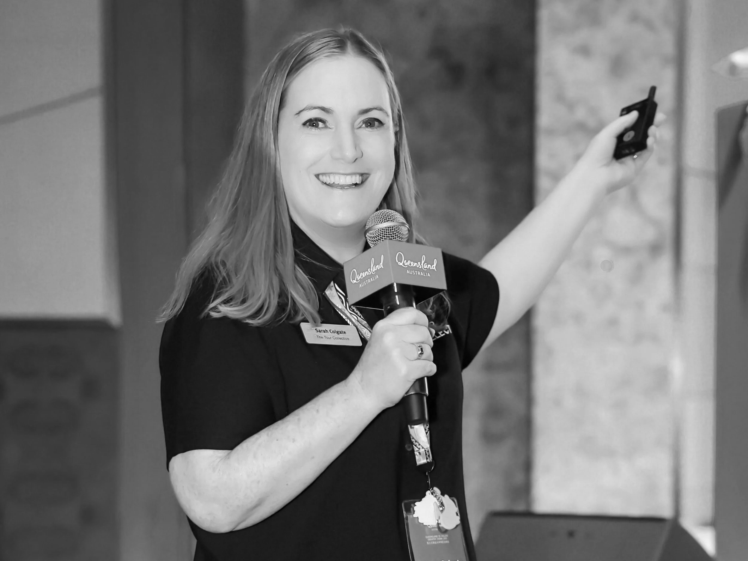 A smiling woman with shoulder-length hair holding a microphone labeled Queensland Australia, and raising her other hand while standing indoors.