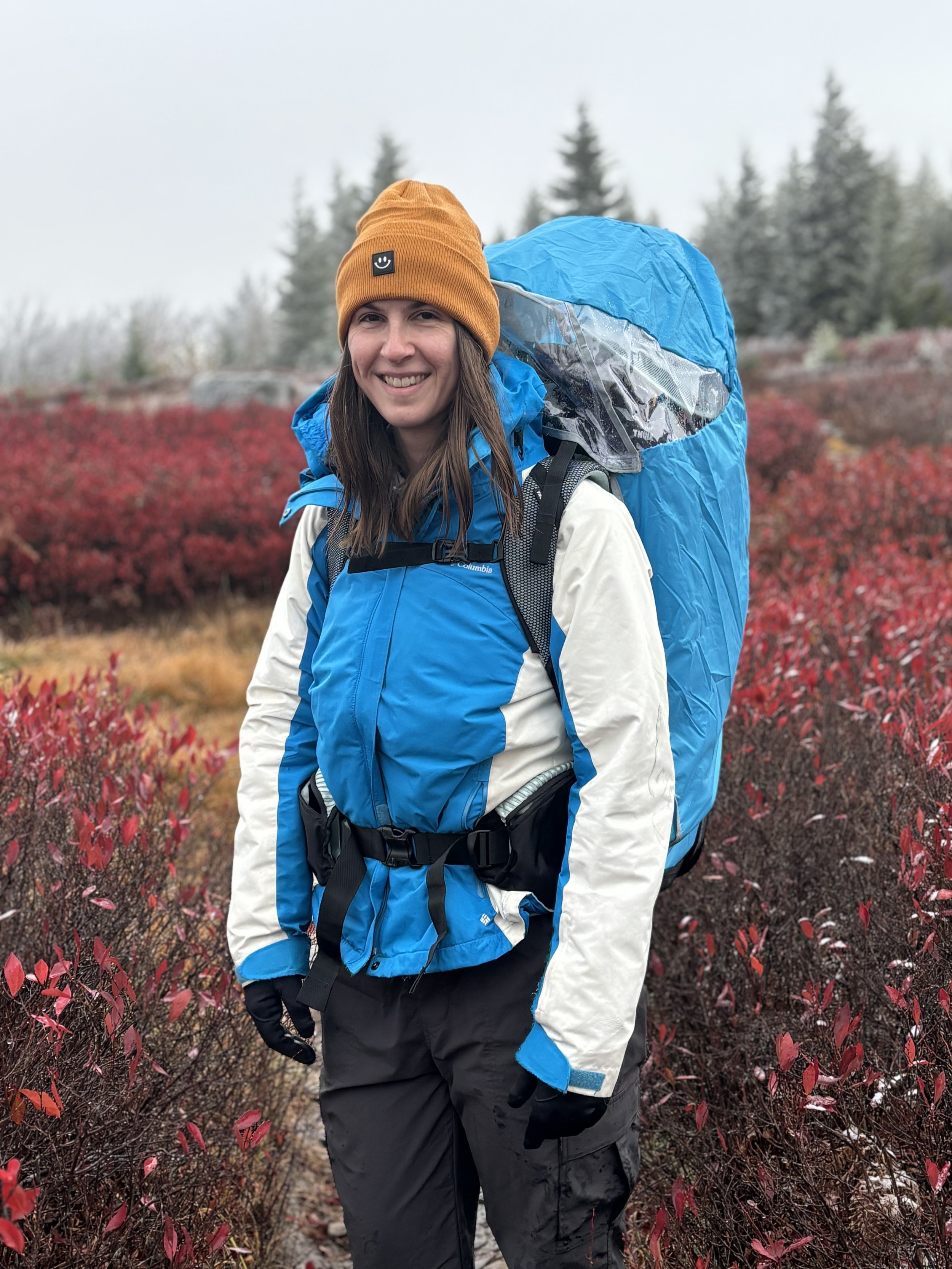 A woman smiling outdoors wearing a blue and white jacket, a mustard beanie, and black gloves, standing on a trail surrounded by red bushes, with fog and evergreen trees in the background.