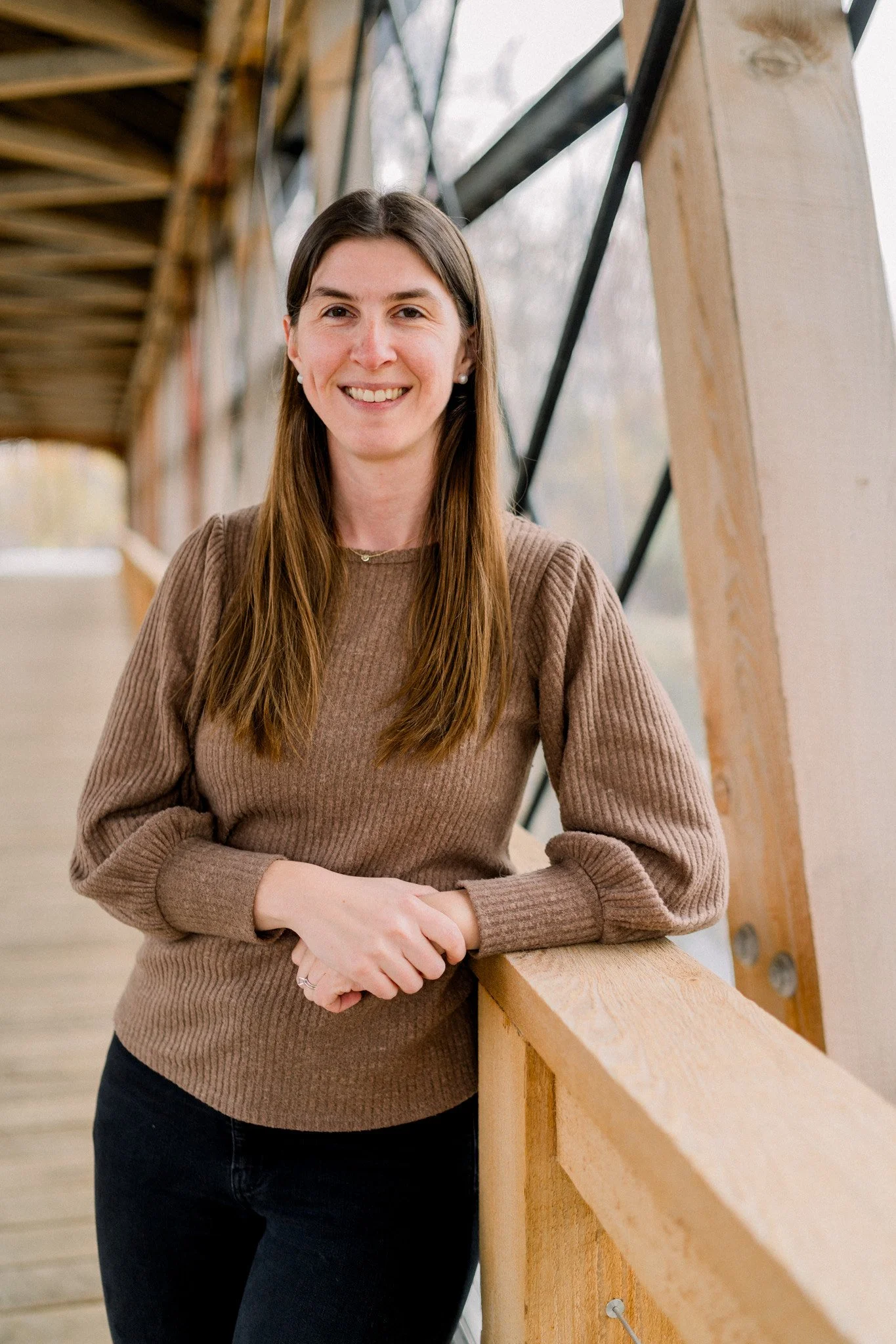 A smiling woman with long brown hair wearing a brown sweater and black pants, leaning on a wooden railing inside a building with large windows and exposed wooden beams.