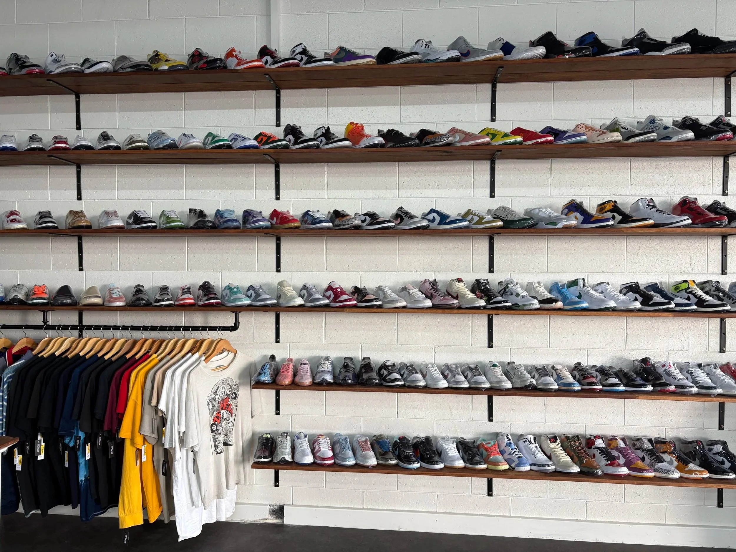 Wall display of various athletic shoes arranged on wooden shelves, with T-shirts hanging on a rack below.