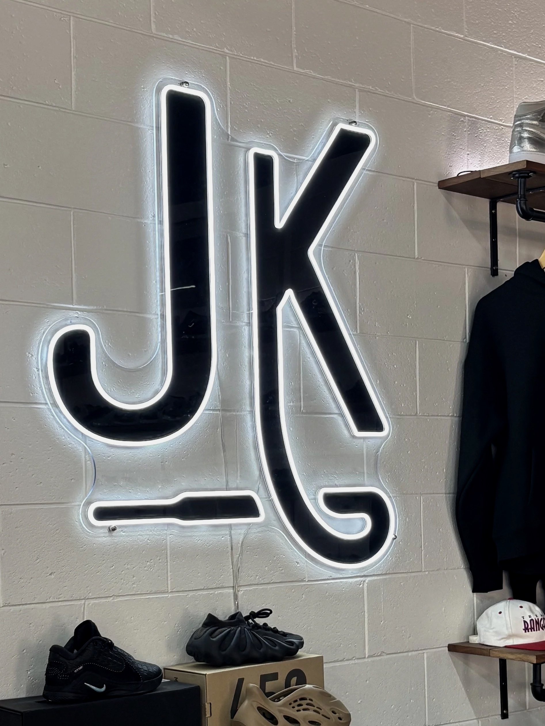 Neon sign with the word 'JLK' in black letters outlined with white light, mounted on a white brick wall inside a retail store.
