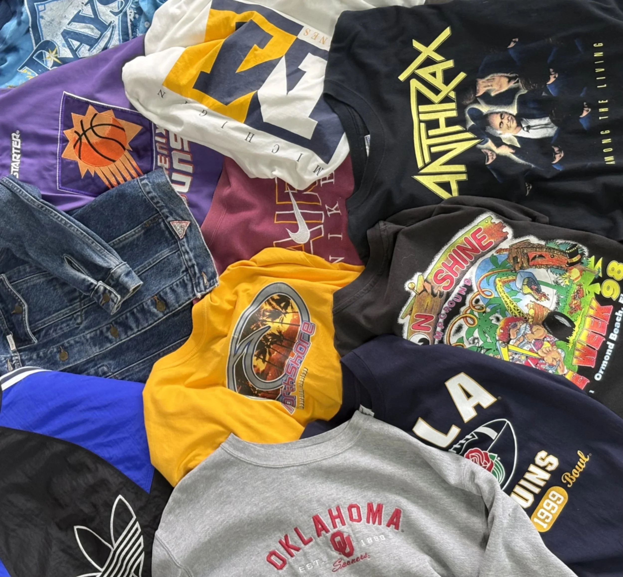 Collection of various vintage and graphic T-shirts, with some folded jeans visible, laid out on a flat surface.