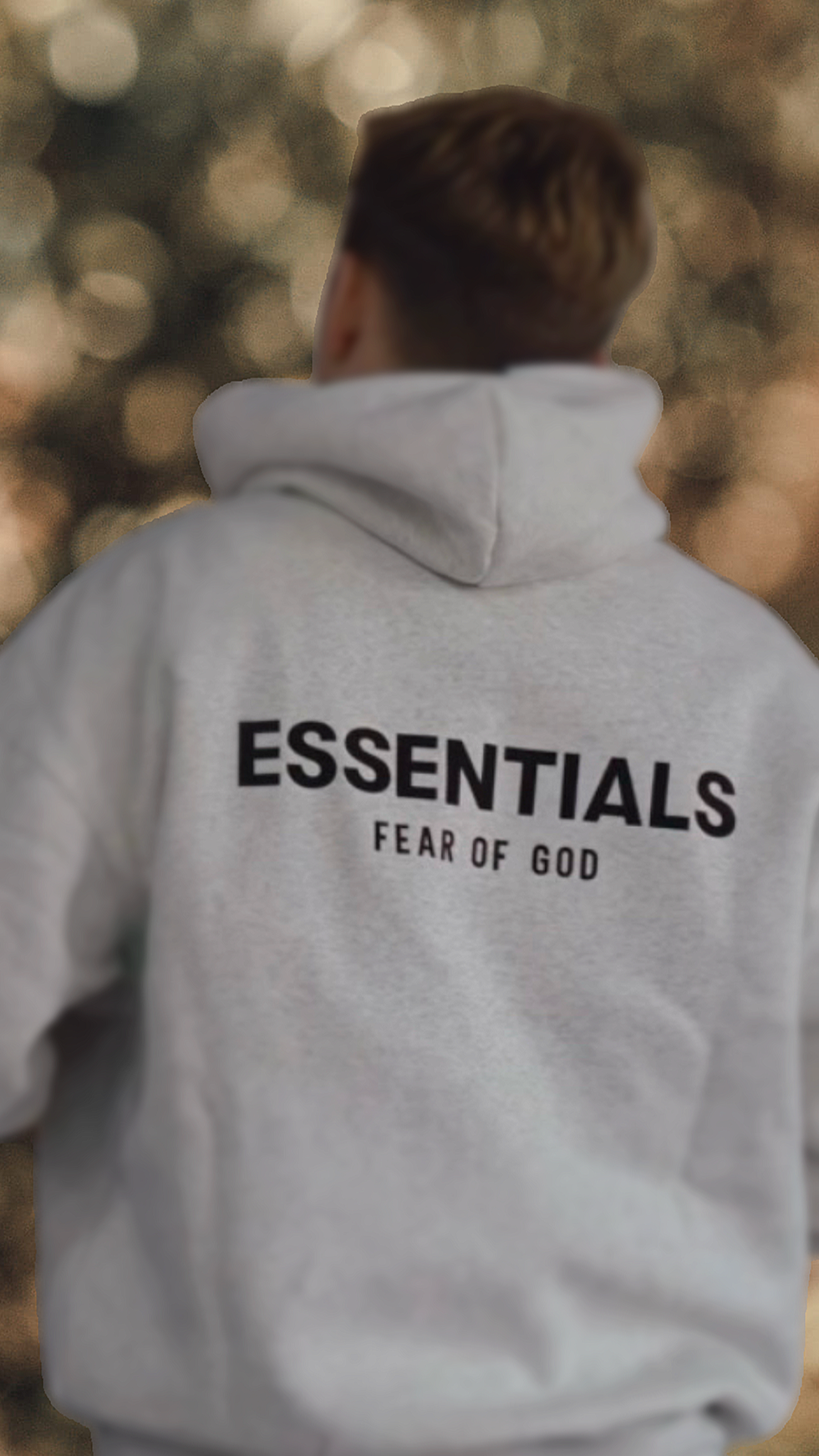 Person wearing a gray hoodie with the words "ESSENTIALS FEAR OF GOD" printed on the back, seen from behind outdoors.