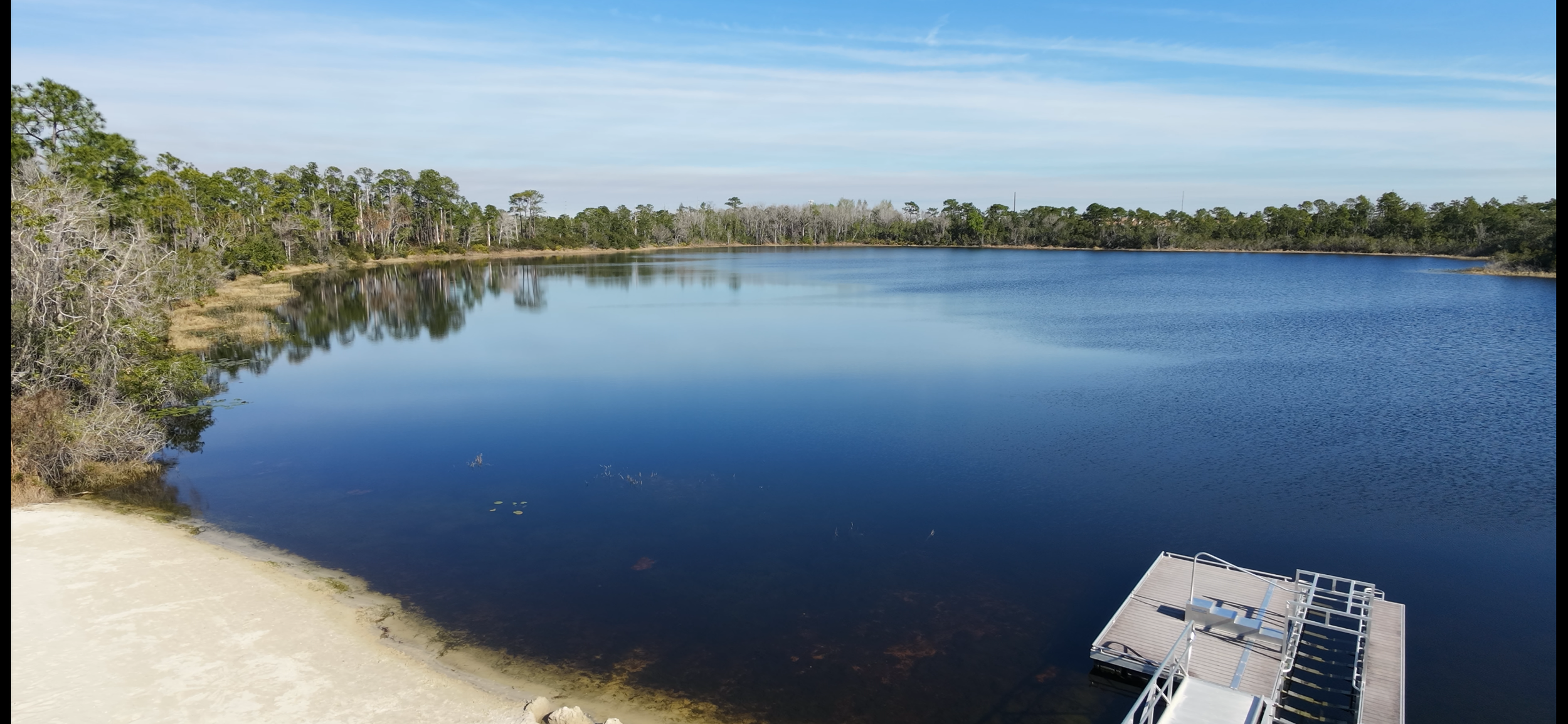 A calm lake surrounded by trees, with a small sandy beach and a dock in the foreground.
