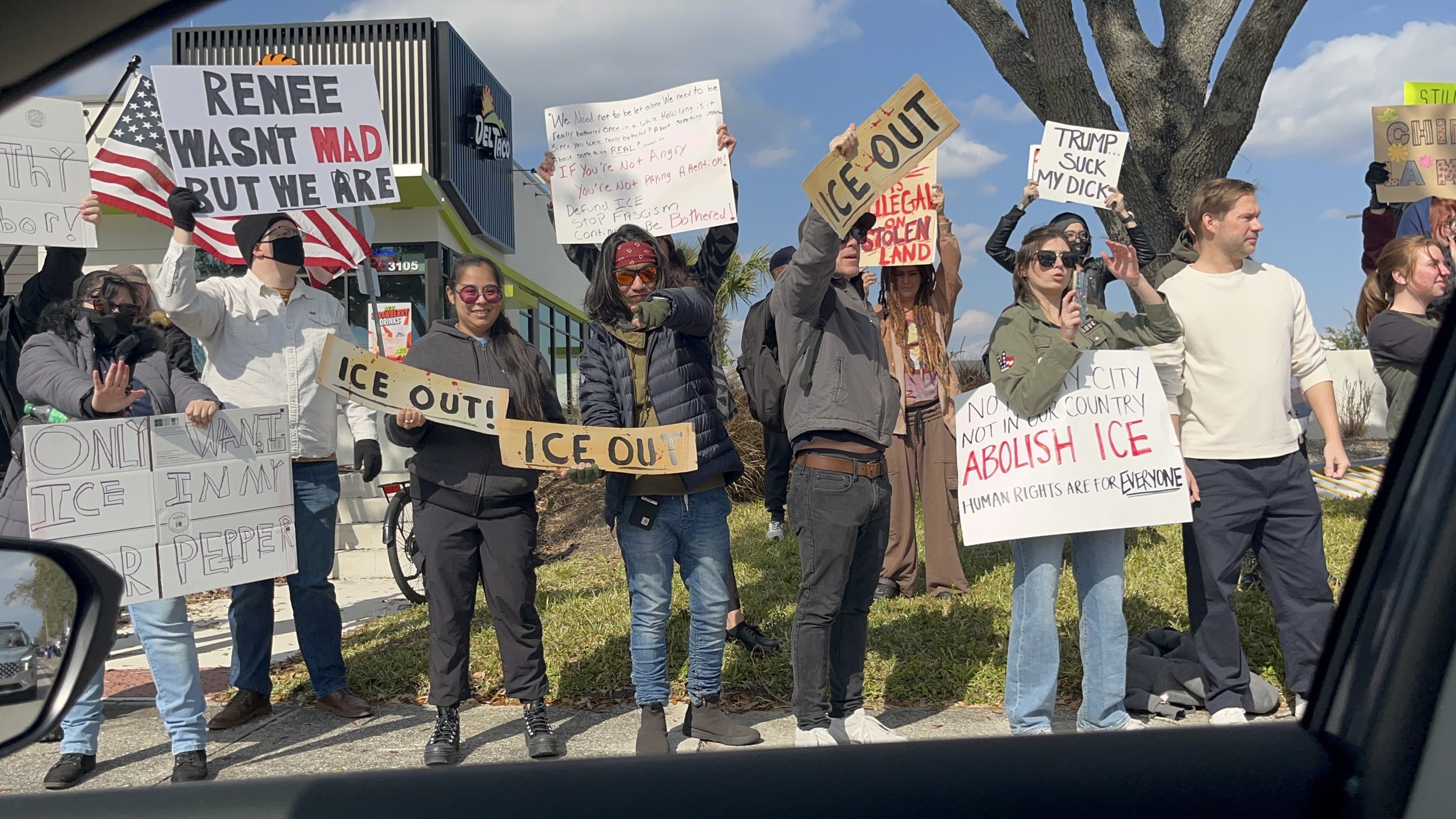 Group of protesters holding signs and banners advocating for the end of ice and immigration enforcement, with some signs reading "ICE OUT," "RENÉE WASN'T MAD BUT WE ARE," "ONLY WANT ICE IN MY RIPPERS," and "ABOLISH ICE." Participants are standing outside near a building with a tree in the background.