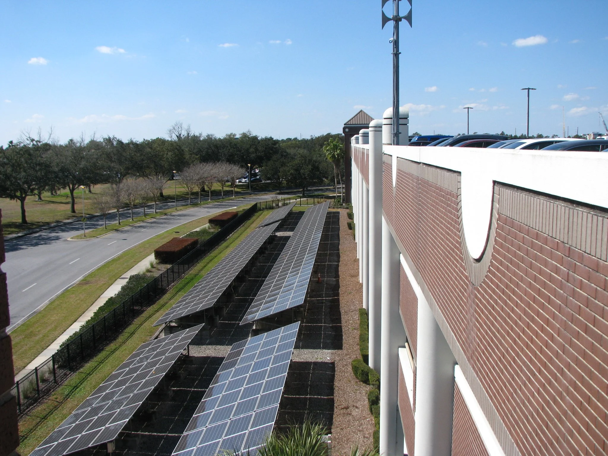 Multiple solar panels installed on the ground outside a building with a brick facade, with a parking lot and trees in the background.