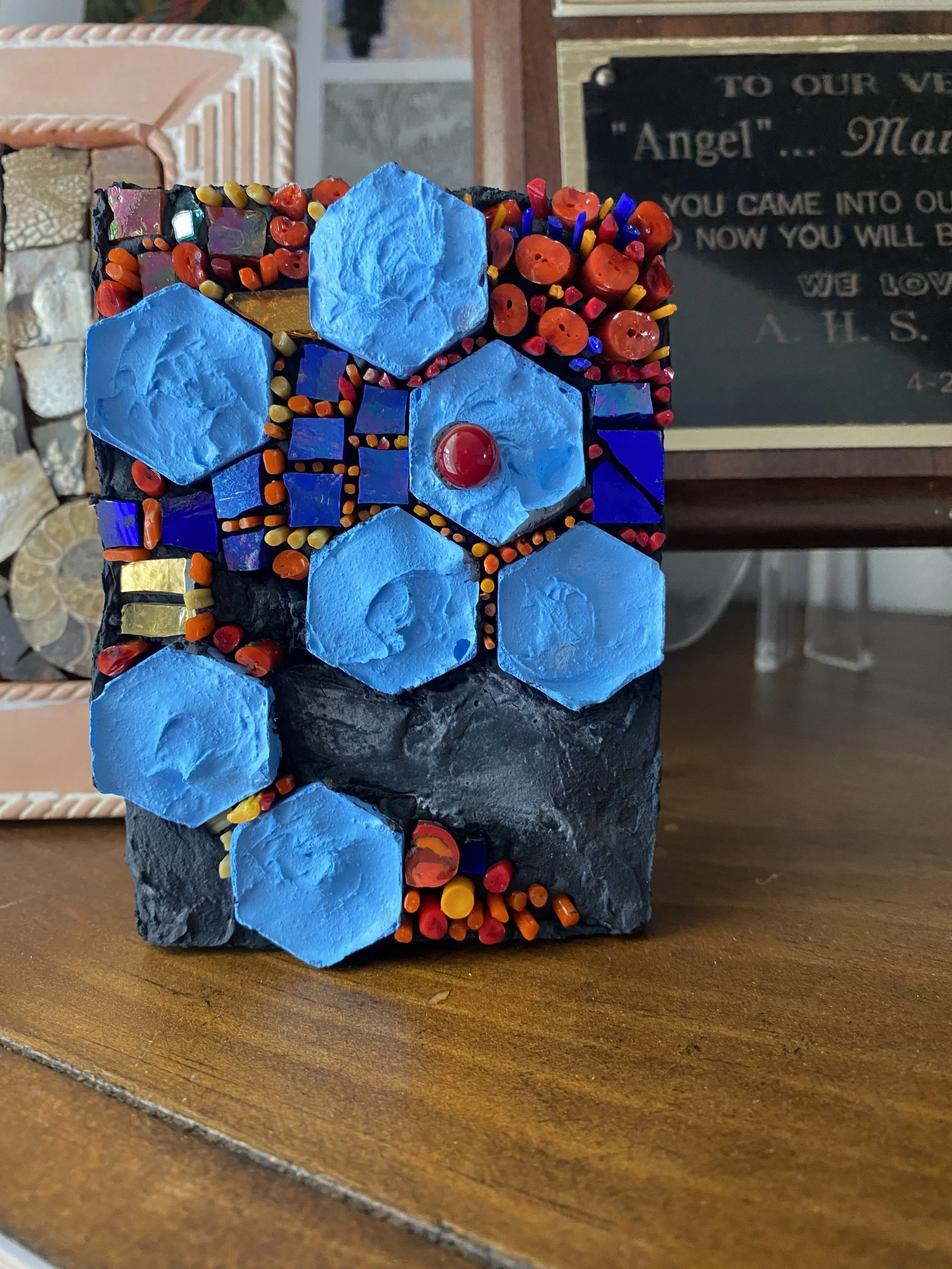 A textured mosaic sculpture featuring large blue hexagons, small colorful beads, and black stone, with a red glass sphere at the center, displayed on a wooden surface.