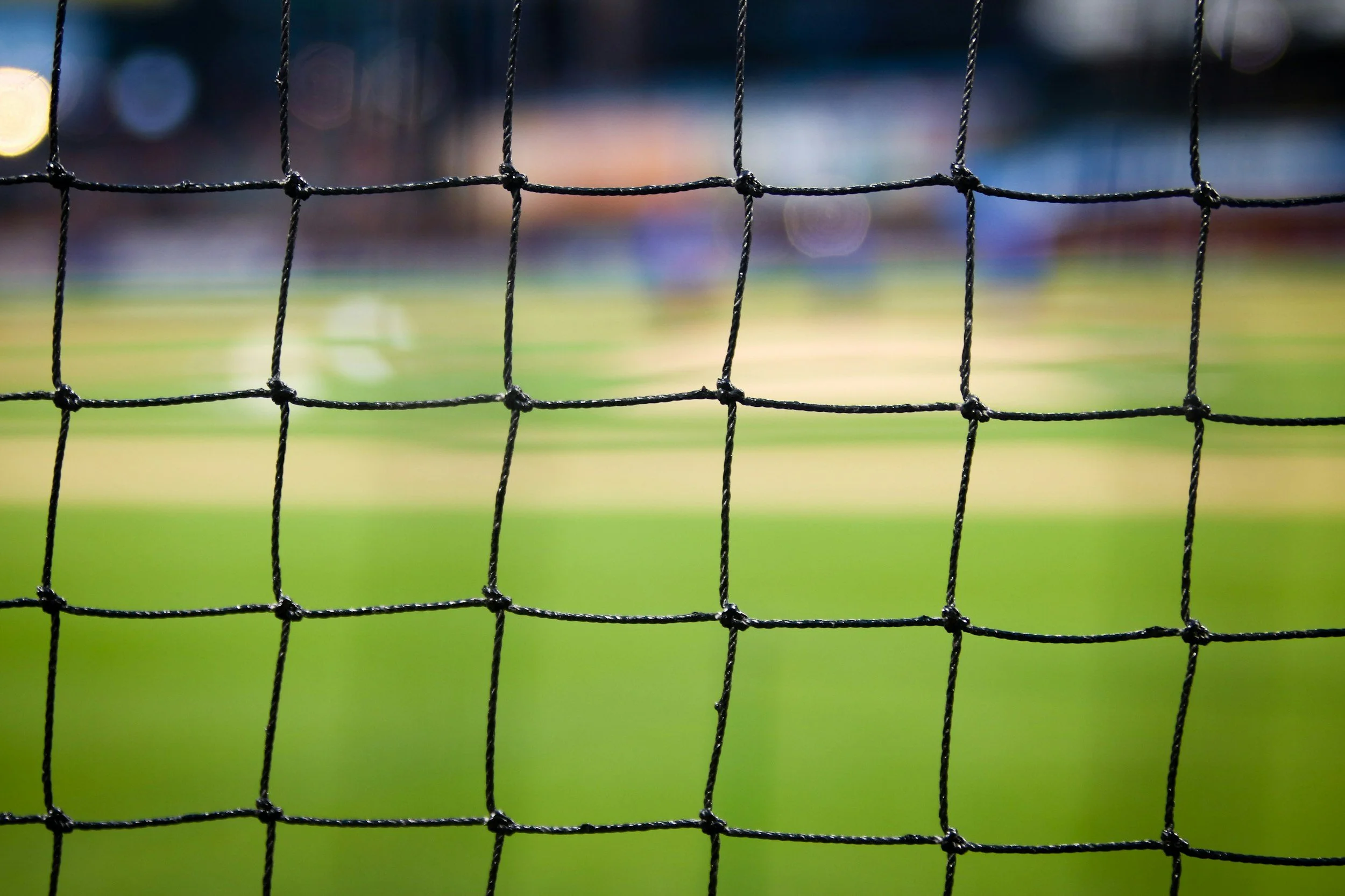 Close-up of a black sports net with a blurred playing field and stadium lights in the background.