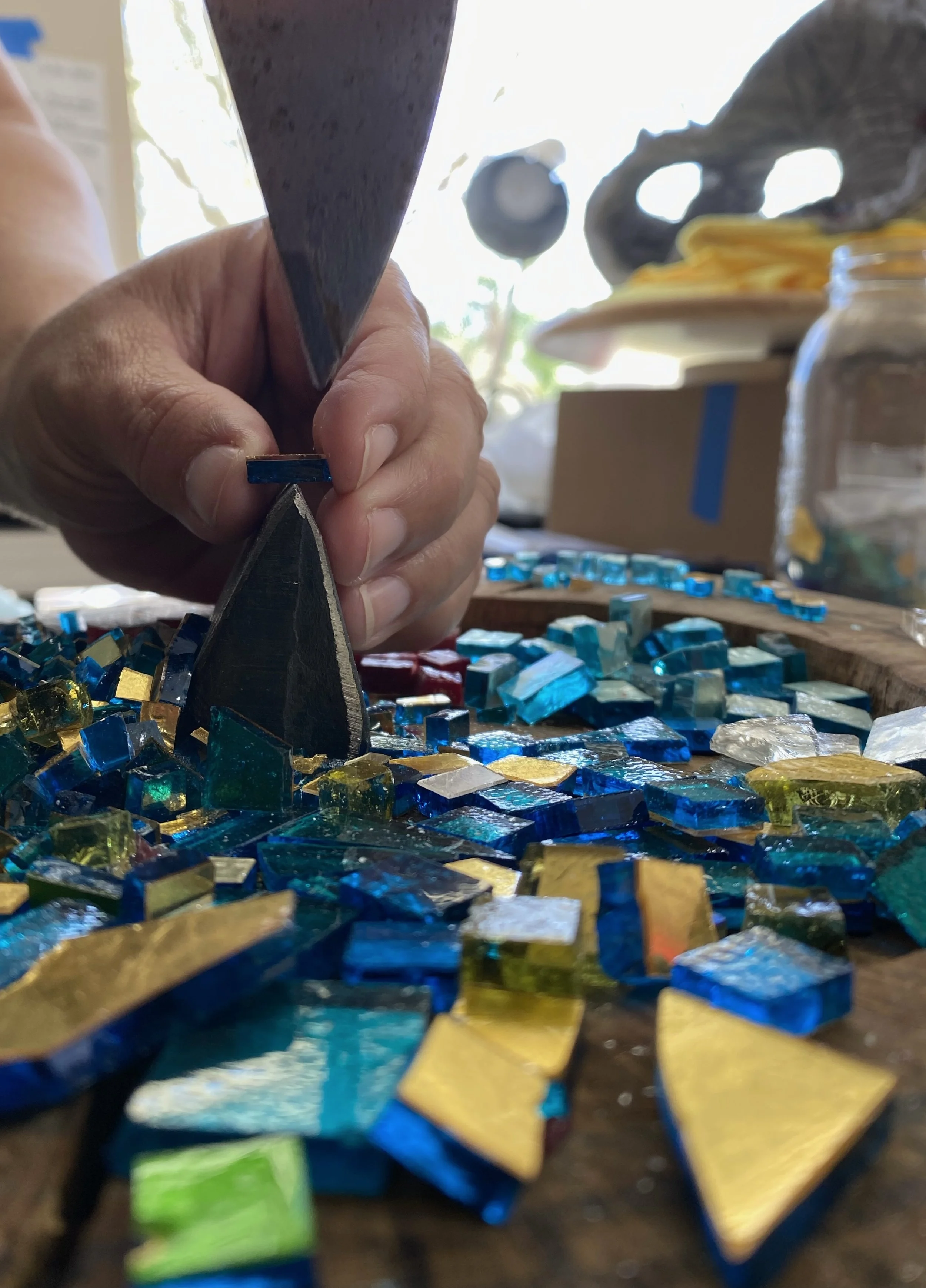 Close-up of a person using a small hammer and chisel to craft colorful glass mosaic tiles on a work surface, with various pieces of glass in blue, green, and gold tones scattered around.