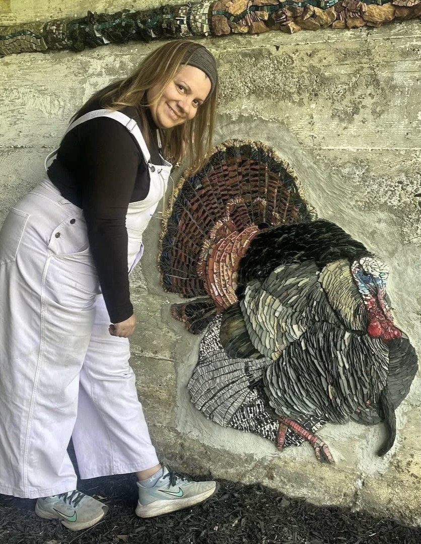 A woman with long blonde hair, wearing a black long sleeve shirt, white overalls, gray Nike sneakers, and a brown headband, smiling while standing next to a mosaic turkey sculpture on a concrete wall.