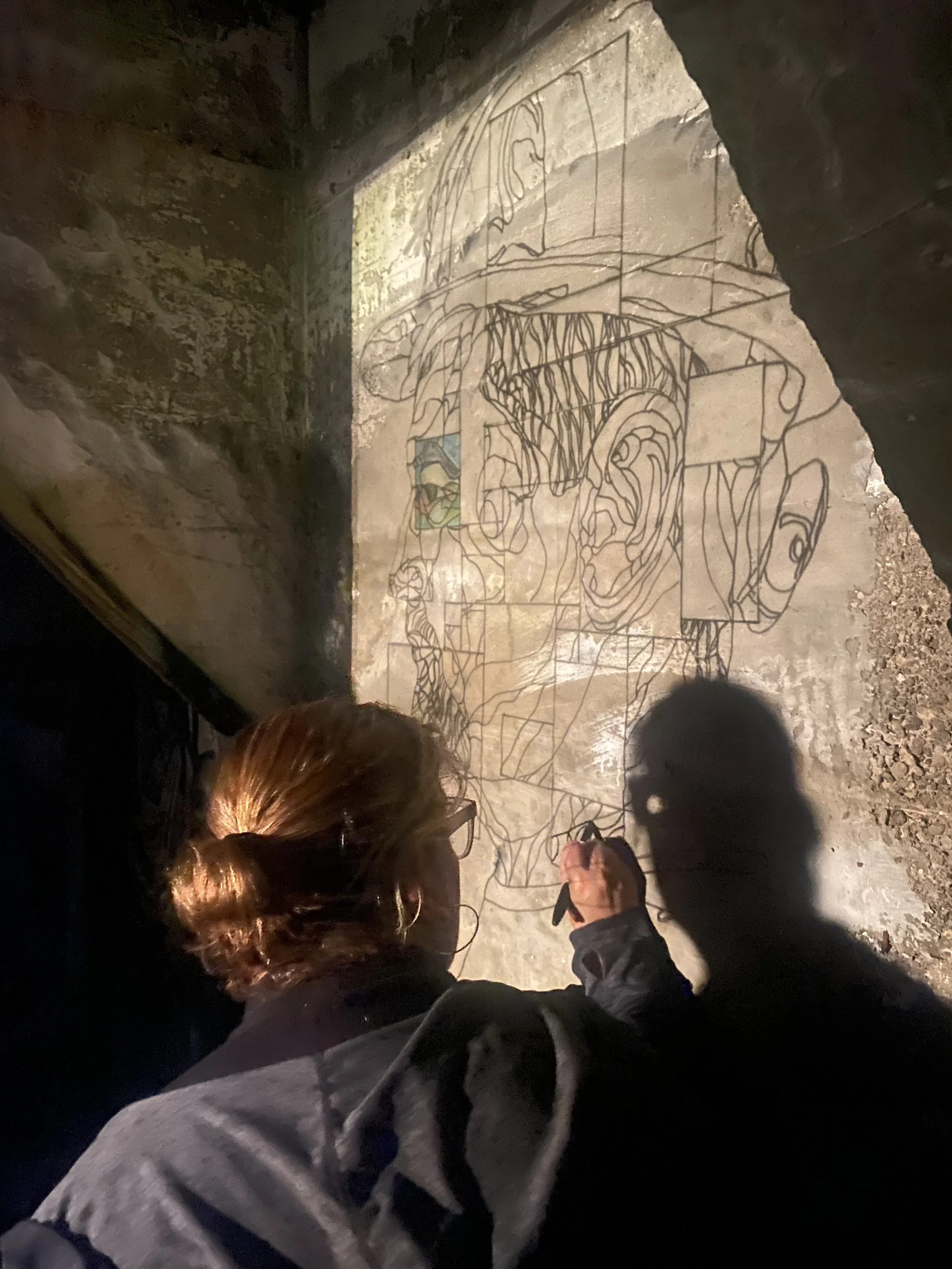A person with reddish-brown hair, wearing glasses, drawing a line art portrait on a concrete wall in a dimly lit space, with a shadow cast on the wall.