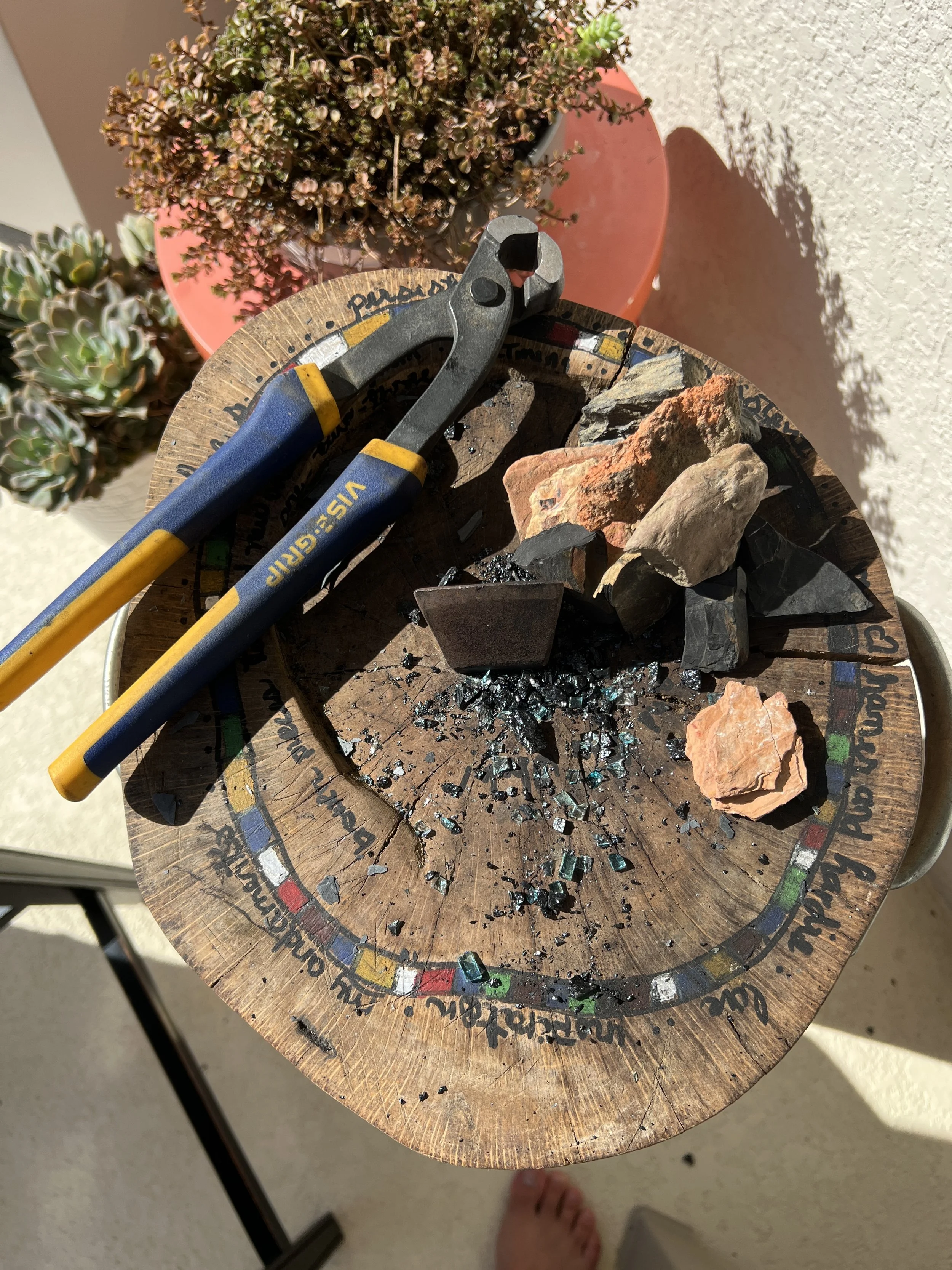 A round wooden table with color-coded letters and text around its edge, a pair of blue and yellow pliers, and various rocks and broken glass. A potted plant with pink flowers is in the background.