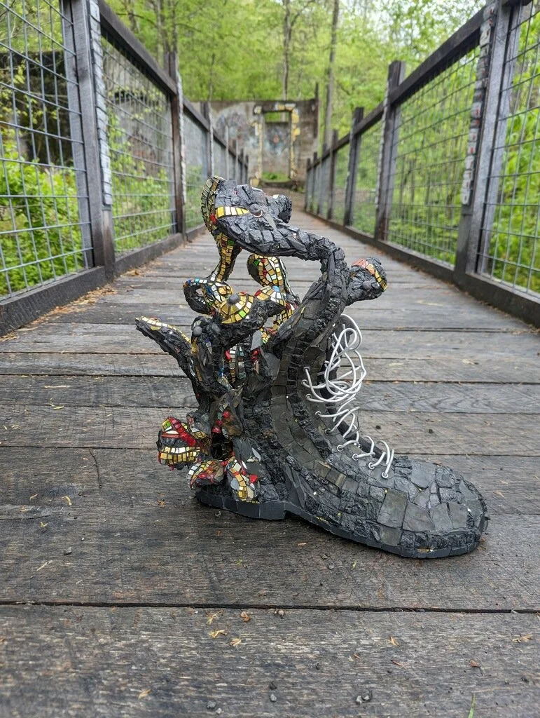 A shoe sculpture made of black material with colorful mosaic accents and white laces, designed to look like a high-top sneaker, placed on a wooden bridge surrounded by greenery.