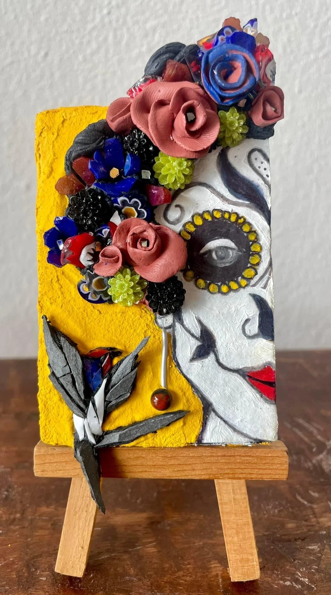 Mixed media artwork featuring a triadic composition with a stylized woman's face, floral arrangements, and painted patterns, displayed on a small wooden easel.