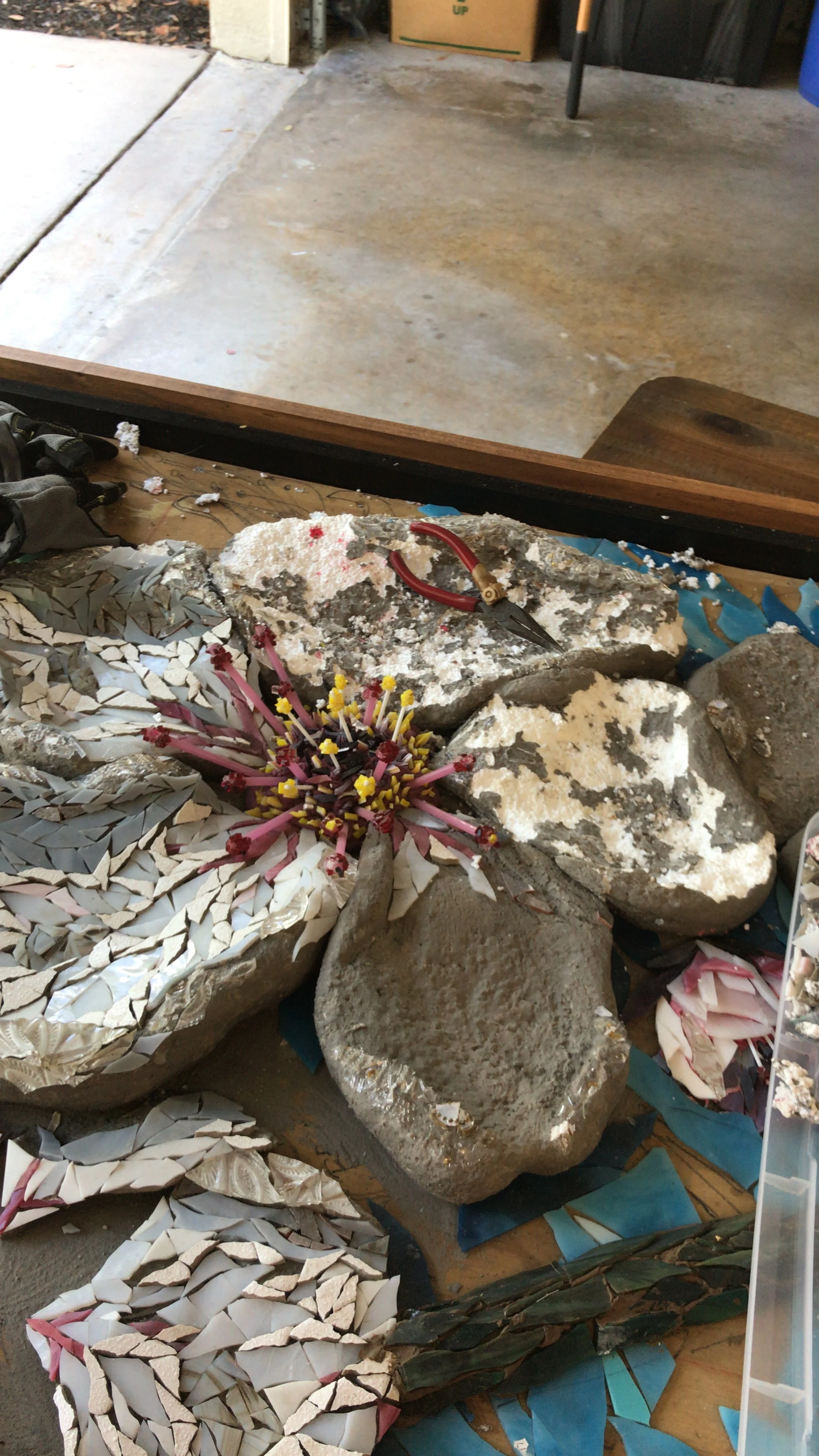 Array of broken tiles, colorful mosaic pieces, and rocks with tools like pliers on a work surface.