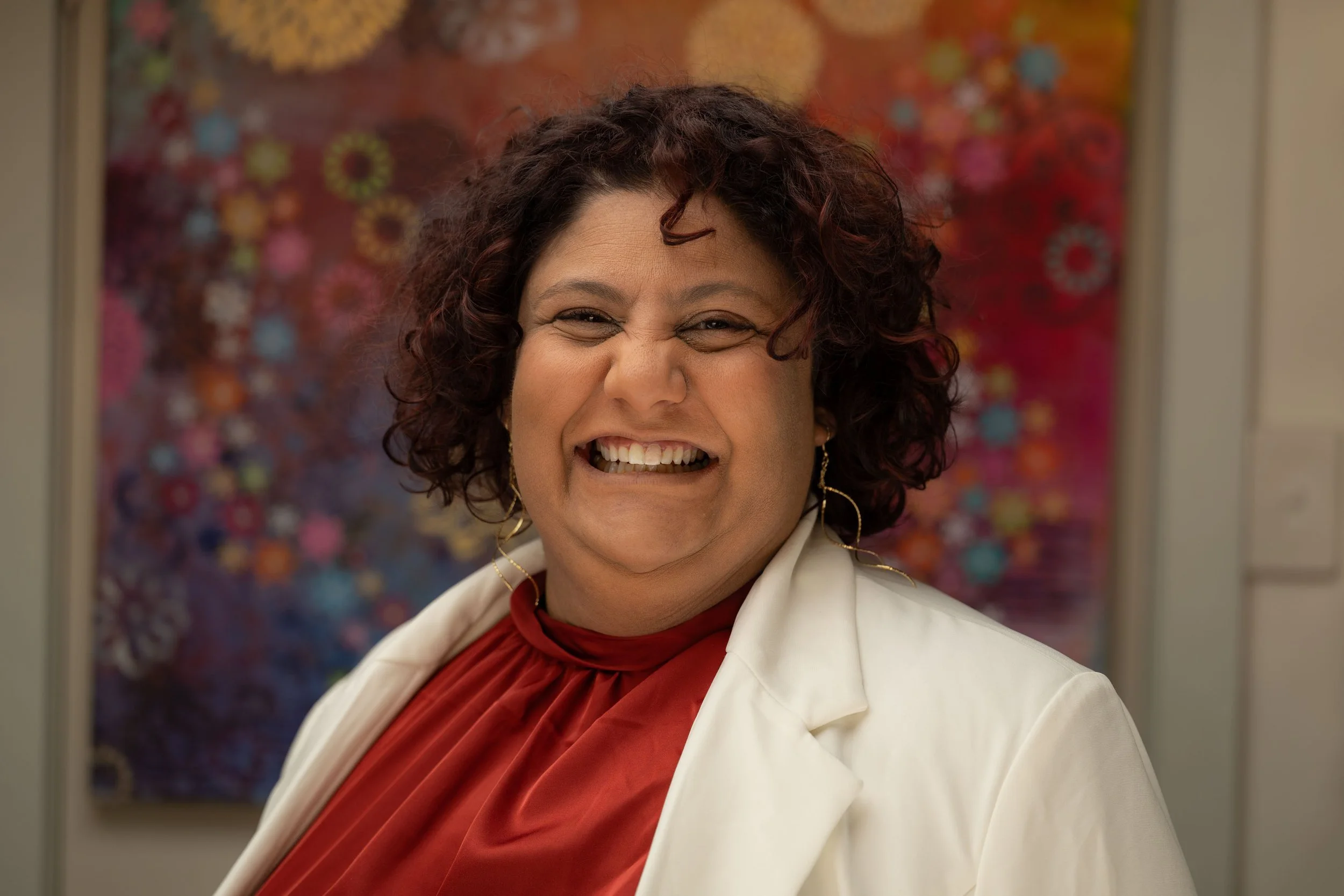 A woman with curly dark hair smiling broadly, wearing a white blazer and red top, with multicolored abstract art in the background.