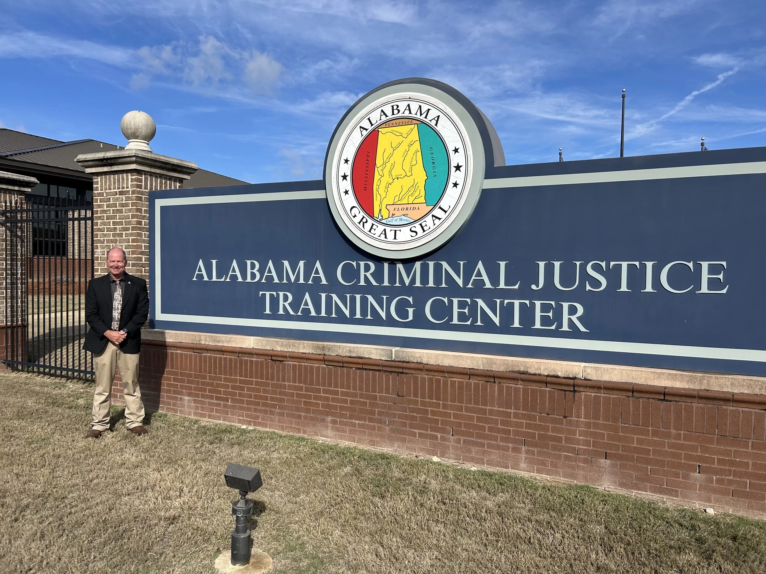 A man wearing a black blazer and beige pants standing next to a large sign that reads 'Alabama Criminal Justice Training Center.' The sign features the official Alabama state seal with a map and the words 'Great Seal' and abbreviations of neighboring states. The background shows a brick building, a blue sky with some clouds, and outdoor lighting fixtures.