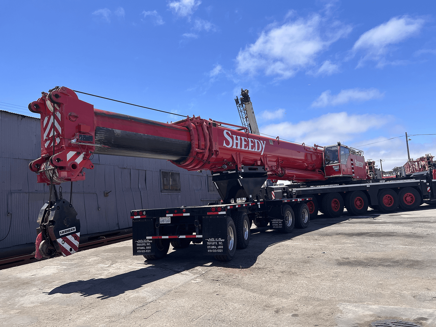 A large red crane truck with the brand name 'SHEEDY' printed on its side, parked on a concrete lot under a blue sky with some clouds.