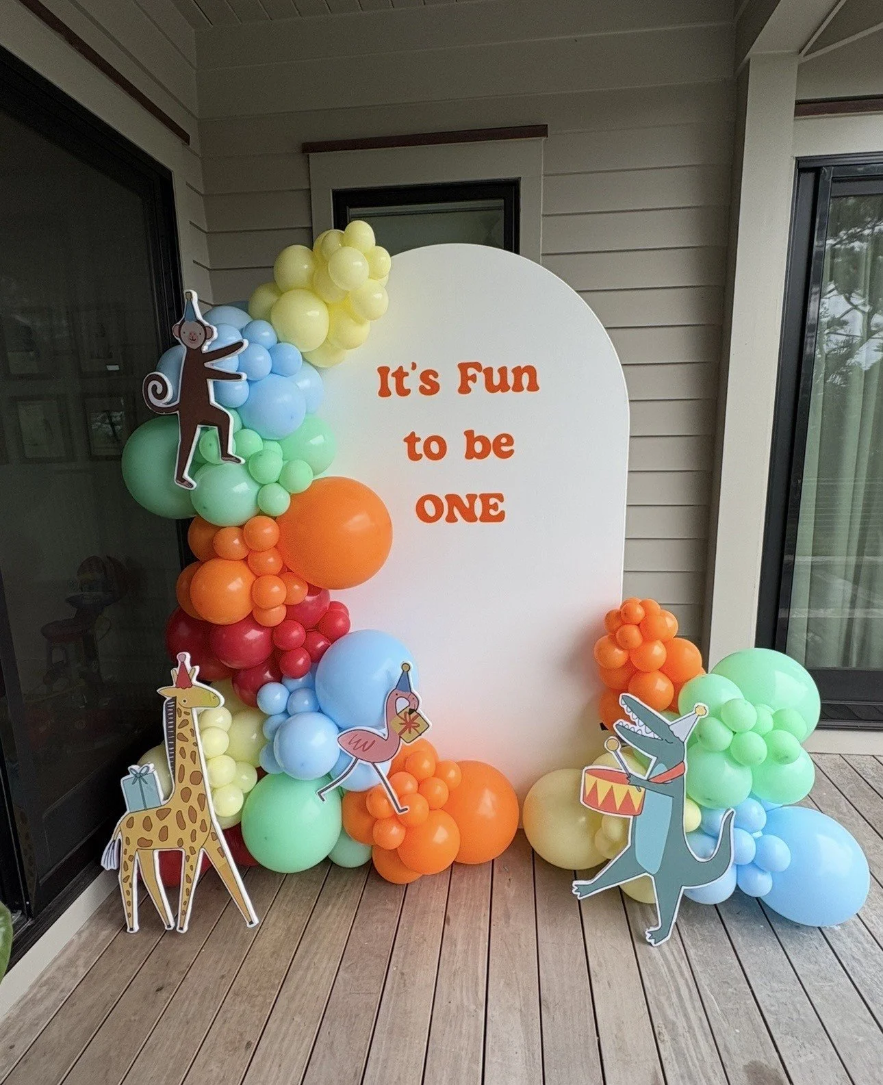 Rainbow colored balloon garland around a white arch backdrop with fun animal cut outs with party hats for a Fun to be One themed 1st Birthday 