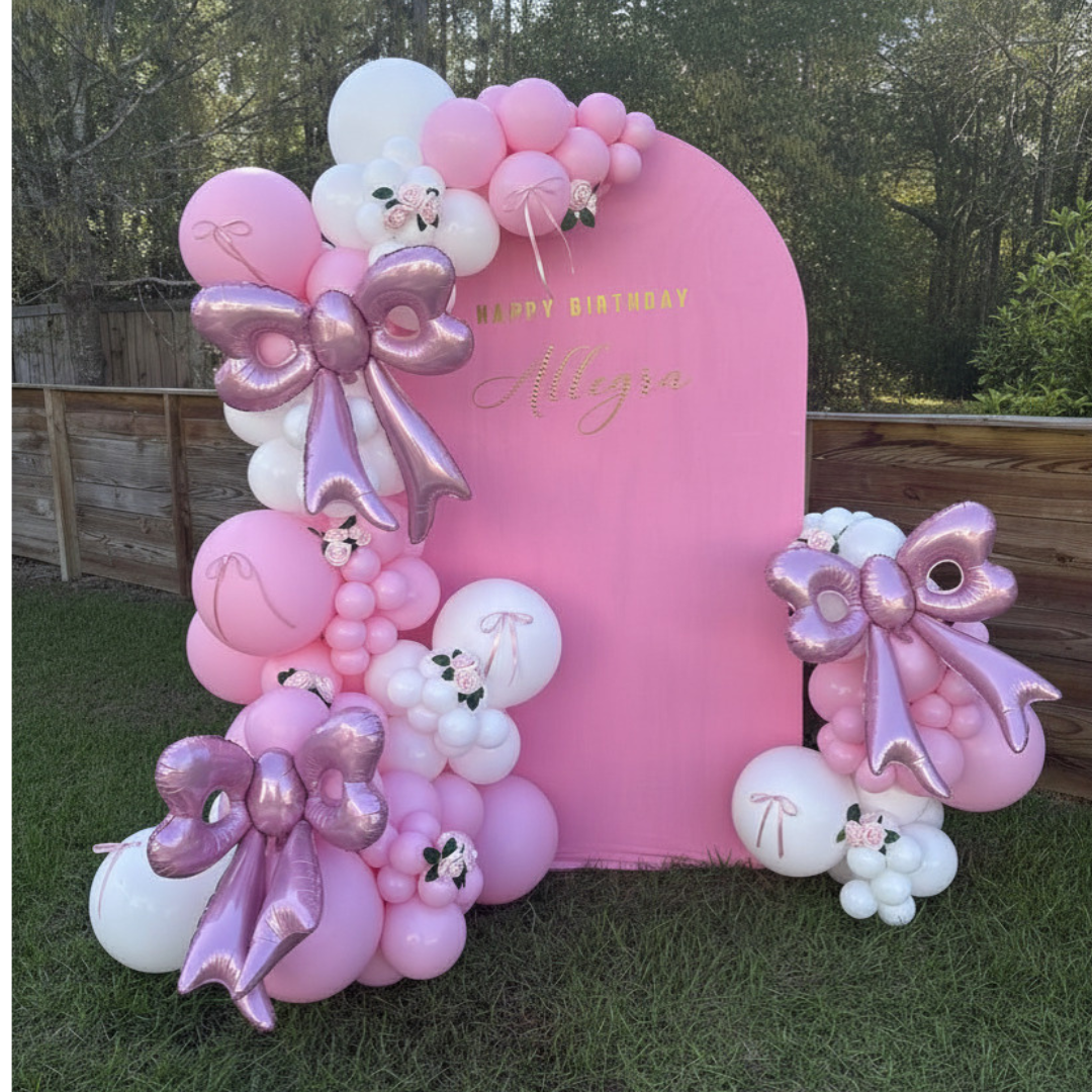 A pink wooden arch backdrop with a pink and white balloon garland with accenting pink ribbon bows and oversized pink mylar balloons and accents of pink roses with some greenery. The backdrop is set in a backyard in Mount Pleasant, SC