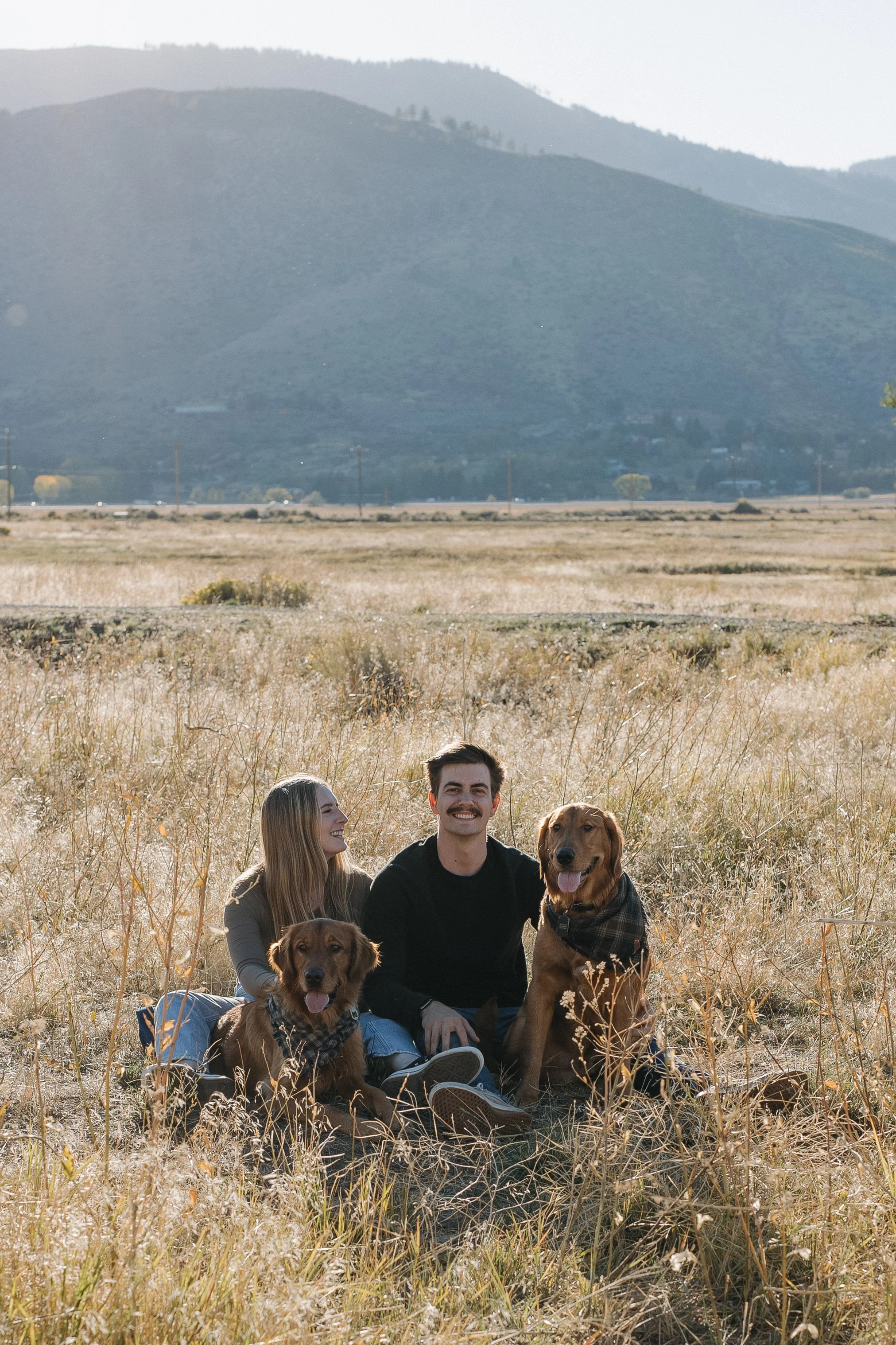 A young couple with two dogs sitting in a grassy field in Washoe Valley with rolling hills in the background.