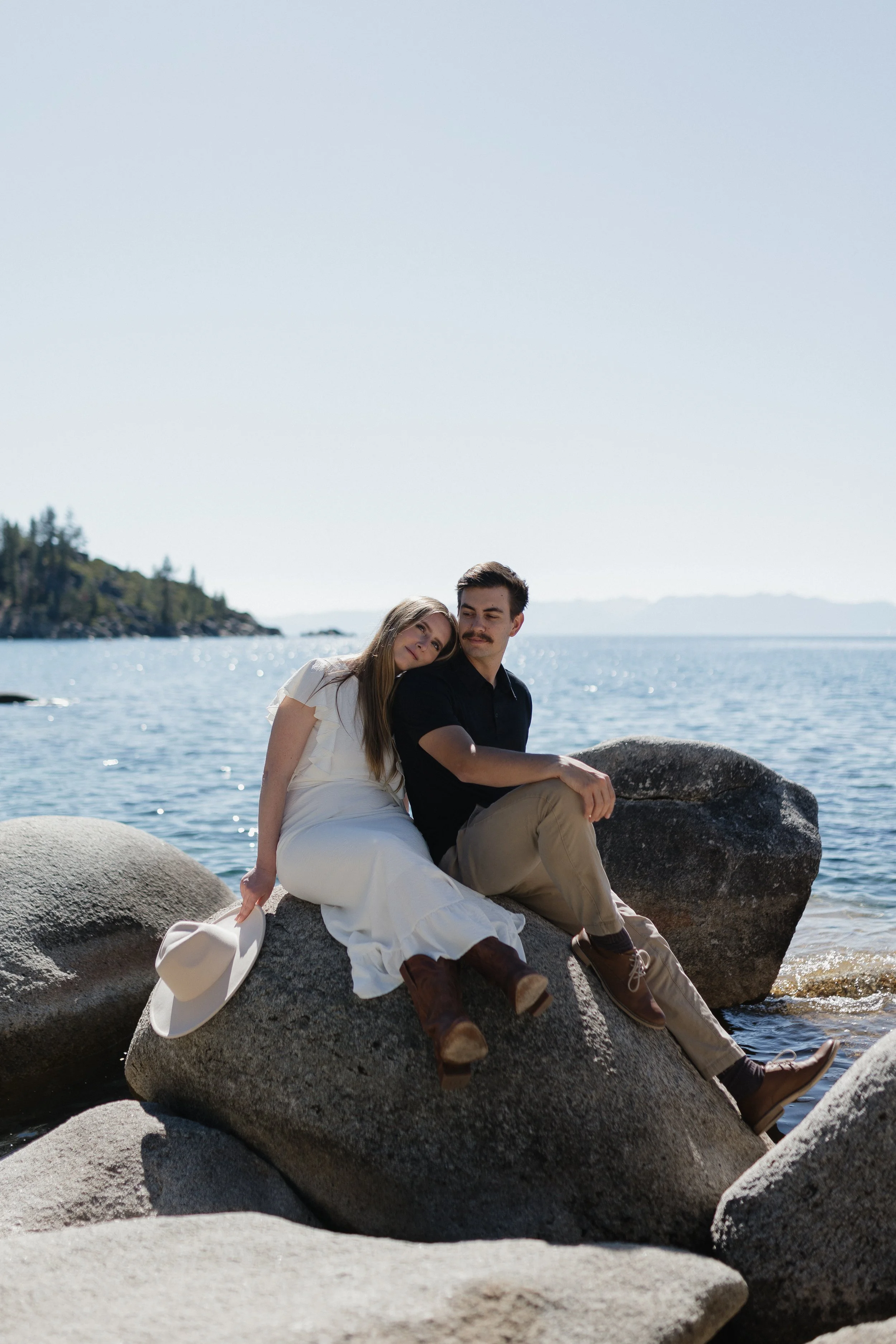 A couple sits on a large rock at Sand Harbor beach in Lake Tahoe by the water near the shoreline, with a scenic view of the lake and distant land in the background, on a bright sunny day.