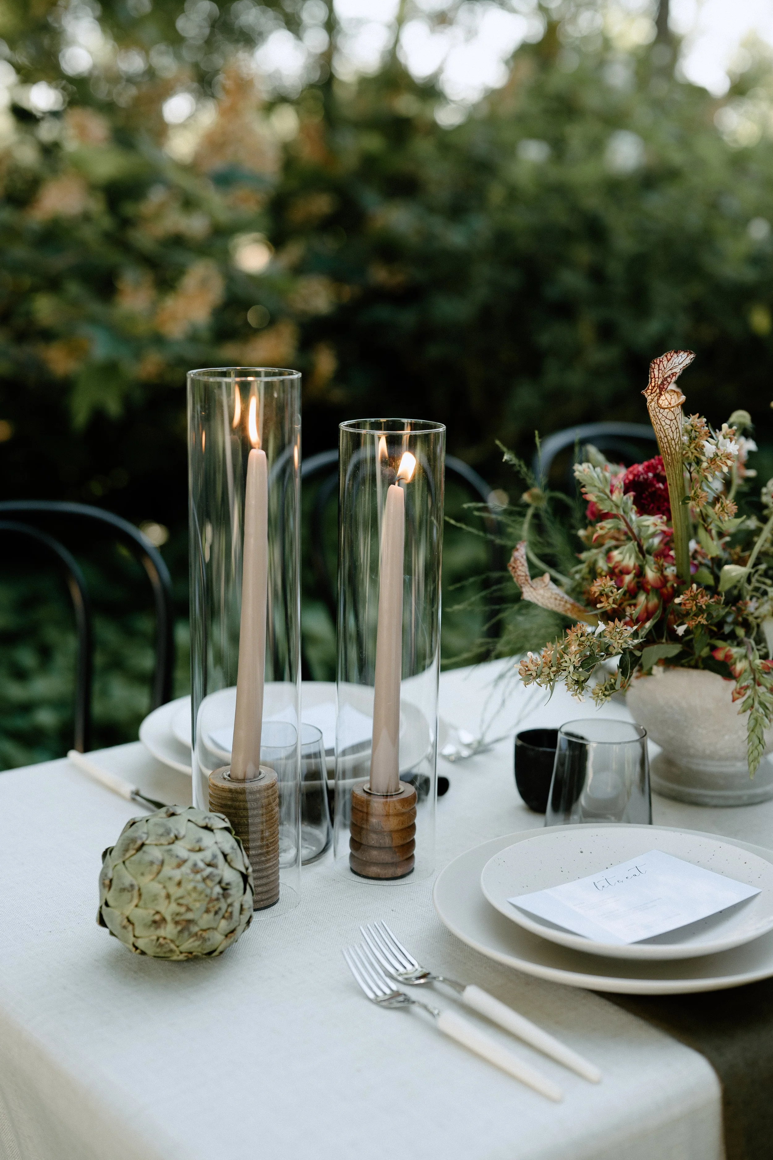Table set outdoors with two tall glass candleholders with lit candles, a white plate with a handwritten note, a fork and knife, a decorative artichoke, a floral arrangement in a white vase, and black and clear drinking glasses.