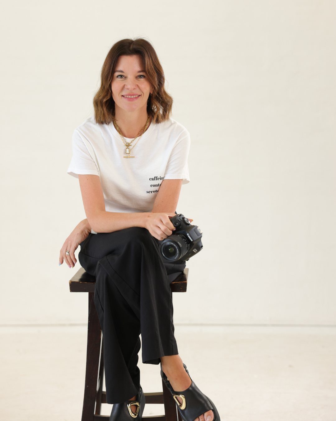 A woman with shoulder-length brown hair sitting on a stool, holding a camera, and smiling at the camera. She is wearing a white t-shirt, black pants, and black shoes, and is accessorized with layered necklaces.