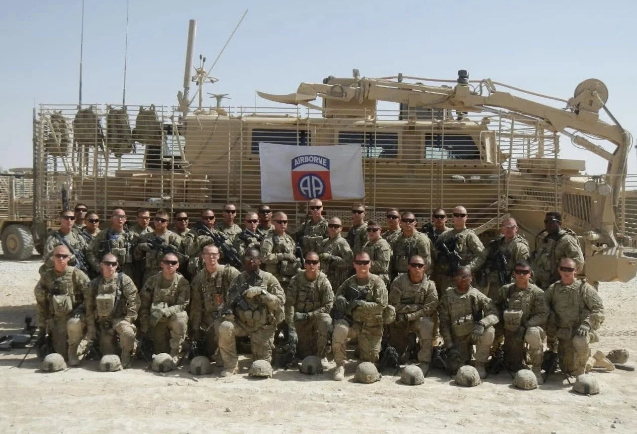 Group of soldiers in combat uniforms posing in front of a military vehicle with a flag that reads 'Airborne' and a logo on it, in a desert setting.