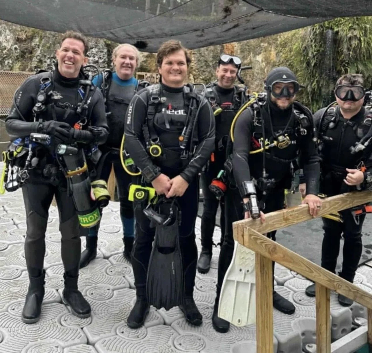 Six scuba divers in black wetsuits and gear, with some wearing goggles and masks, standing on a textured surface near a railing in an outdoor natural setting with trees and rocks.