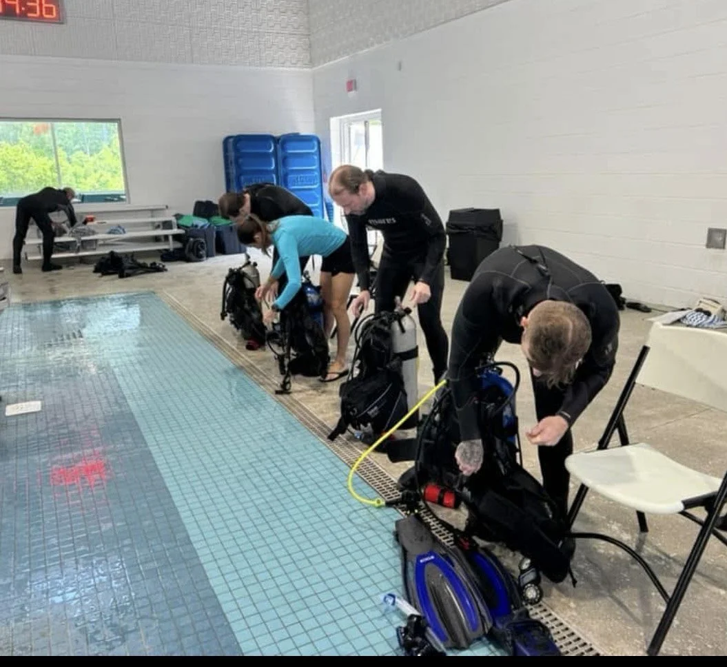 People preparing their diving equipment next to an indoor swimming pool, with gear and bags on the pool deck.
