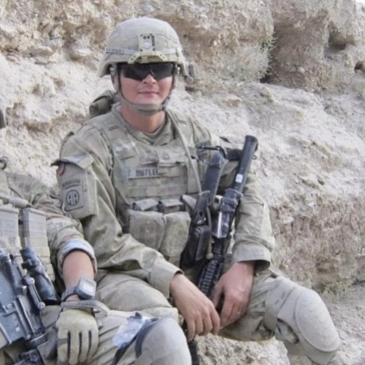 A soldier in camouflage uniform and helmet sitting against a sandy, rocky background, holding a rifle, wearing sunglasses, and smiling.